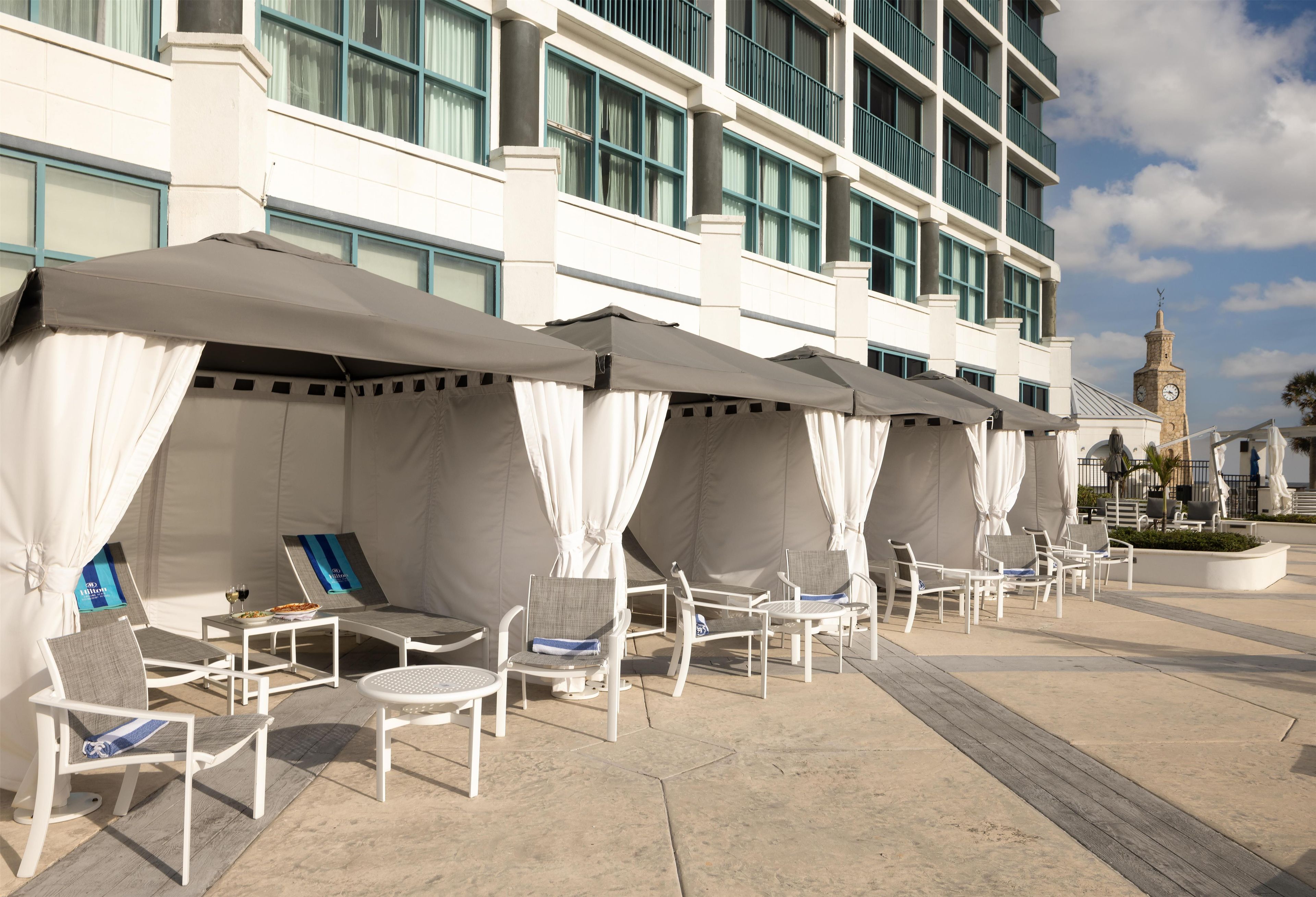 A hotel outdoor seating area with white canopies, lounge chairs, and a clock tower in the background.