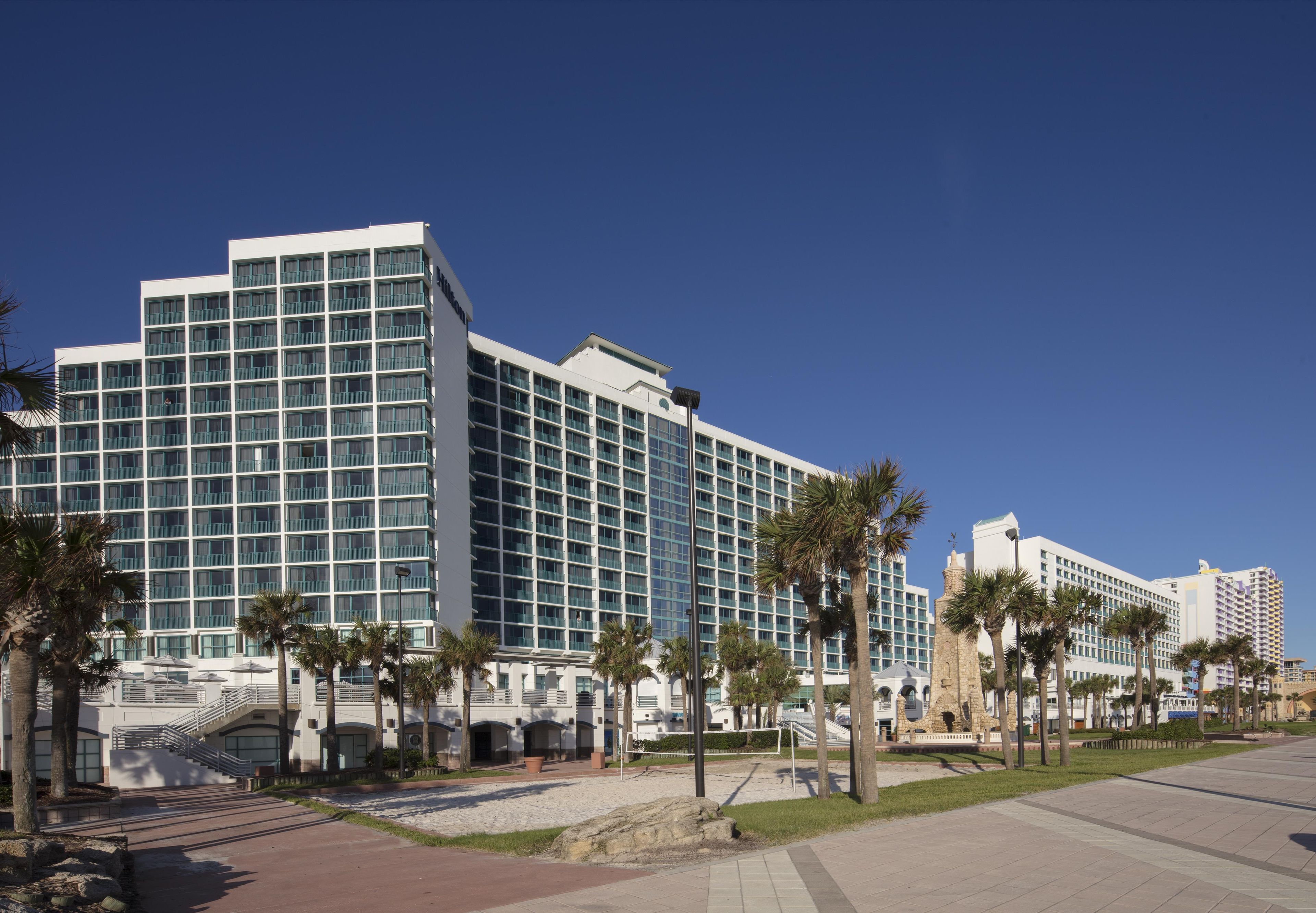 A modern hotel with multiple floors, a central courtyard with palm trees, and a clear blue sky.