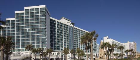 A modern hotel with multiple floors, a central courtyard with palm trees, and a clear blue sky.