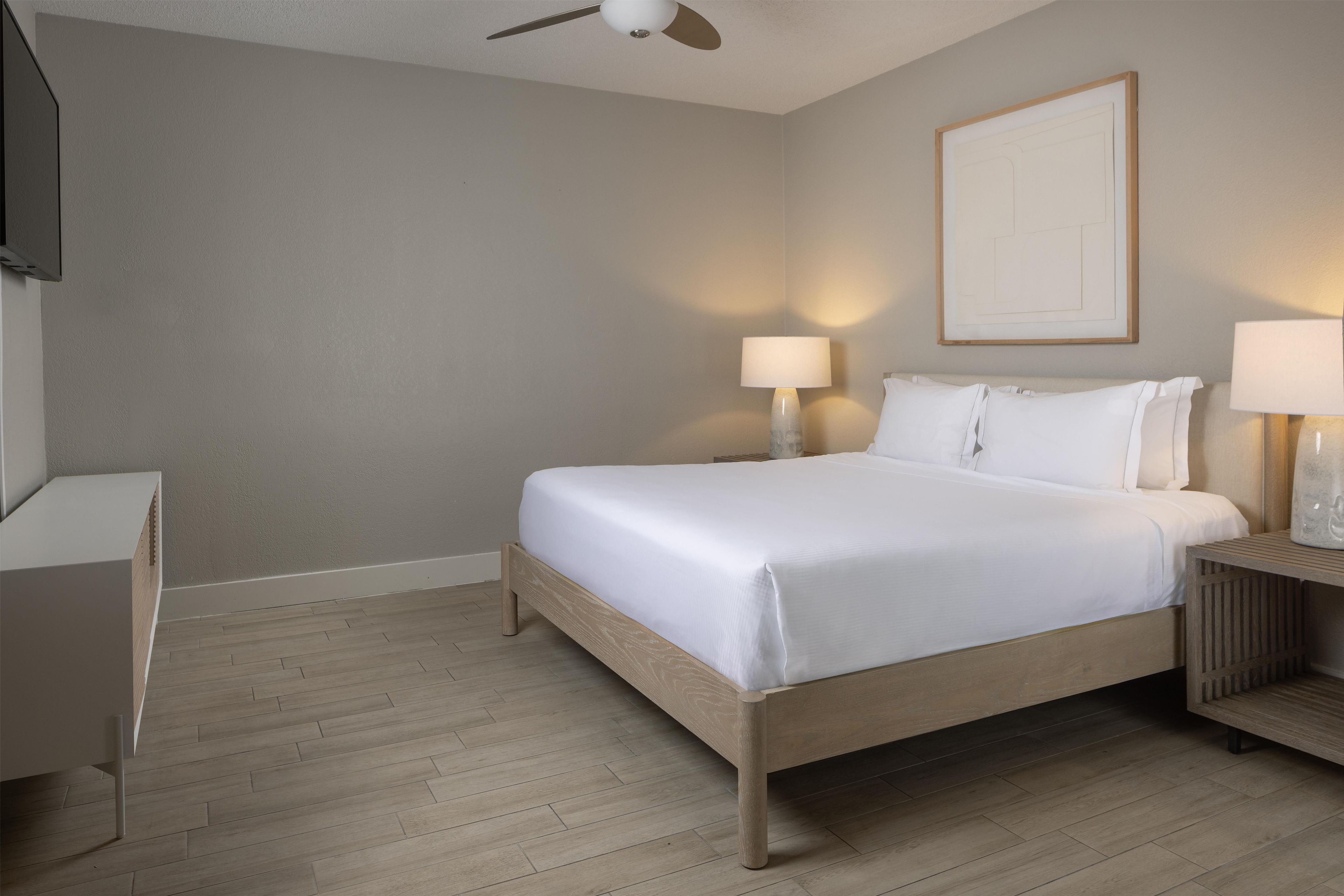 A hotel room with a bed, bedside lamps, a ceiling fan, and a framed picture on the wall.