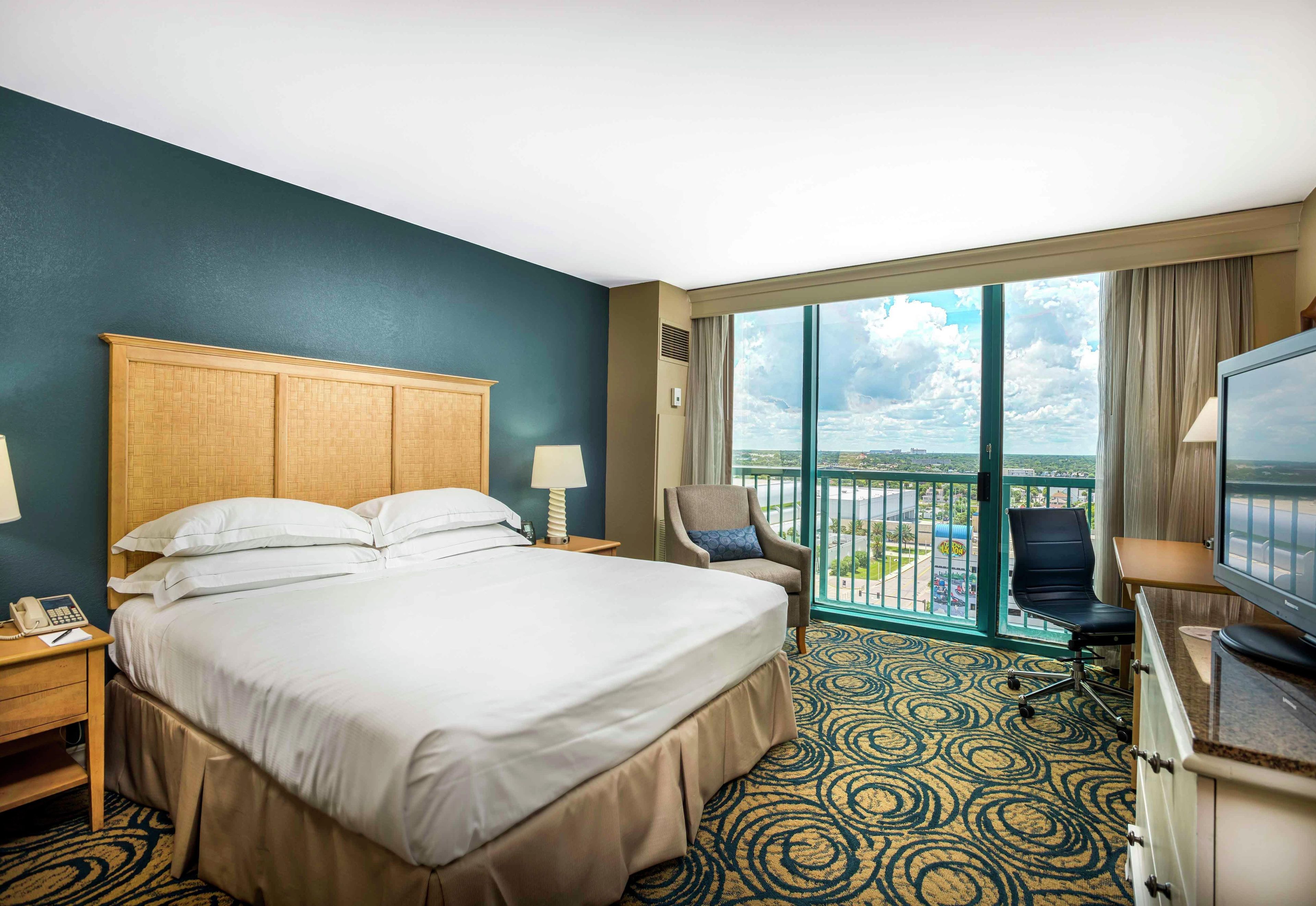Quarto de hotel com cama grande, escrivaninha, cadeira, televisão e varanda com vista.