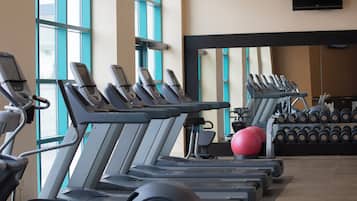 Une rangée de tapis de course, un ballon d’exercice rouge et un porte-haltères dans une salle de sport dotée de grandes fenêtres et d’un téléviseur à écran plat fixé au mur.