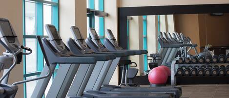 Une rangée de tapis de course, un ballon d’exercice rouge et un porte-haltères dans une salle de sport dotée de grandes fenêtres et d’un téléviseur à écran plat fixé au mur.