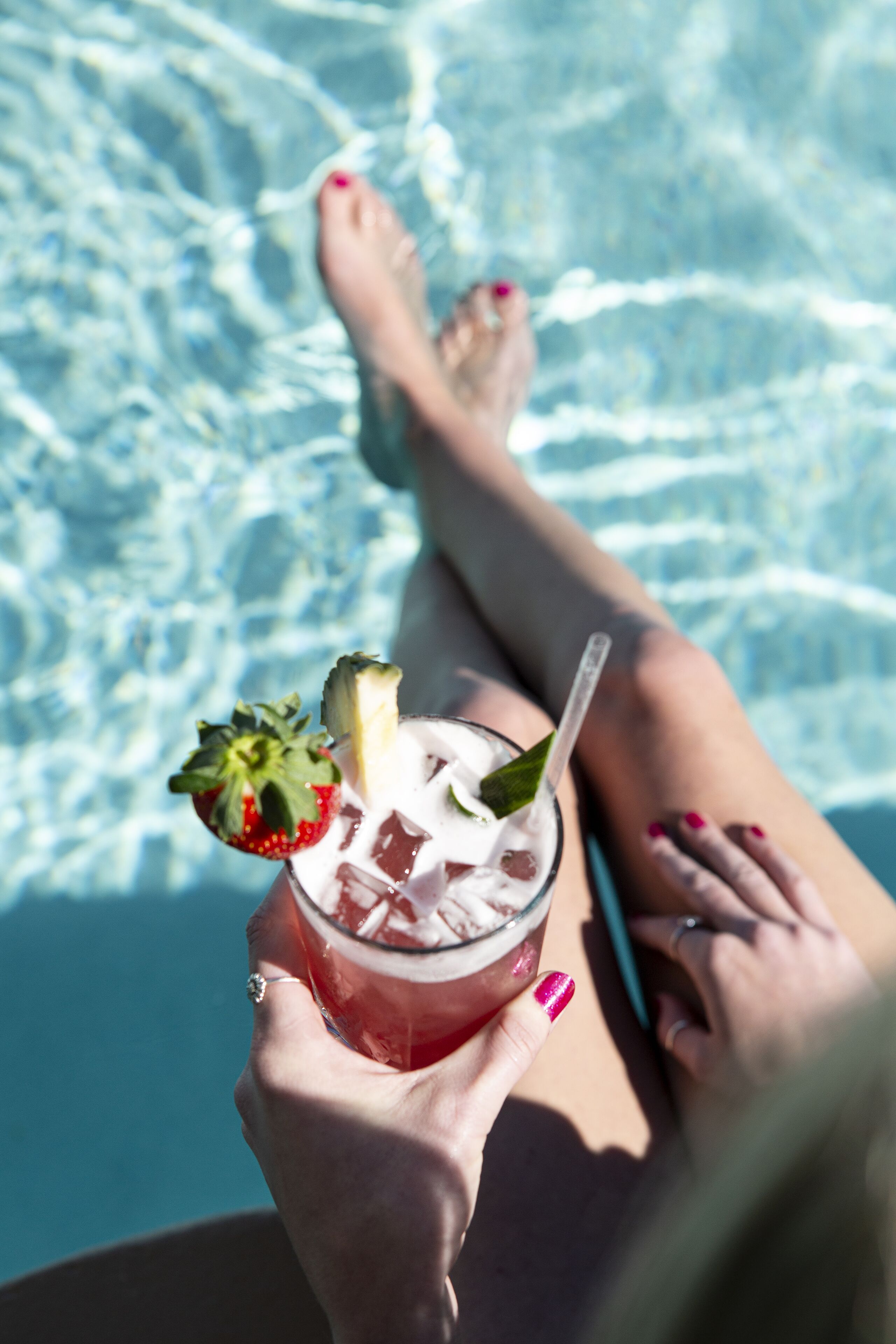 Una persona sosteniendo una bebida con fresa y menta, junto a una piscina de agua cristalina.
