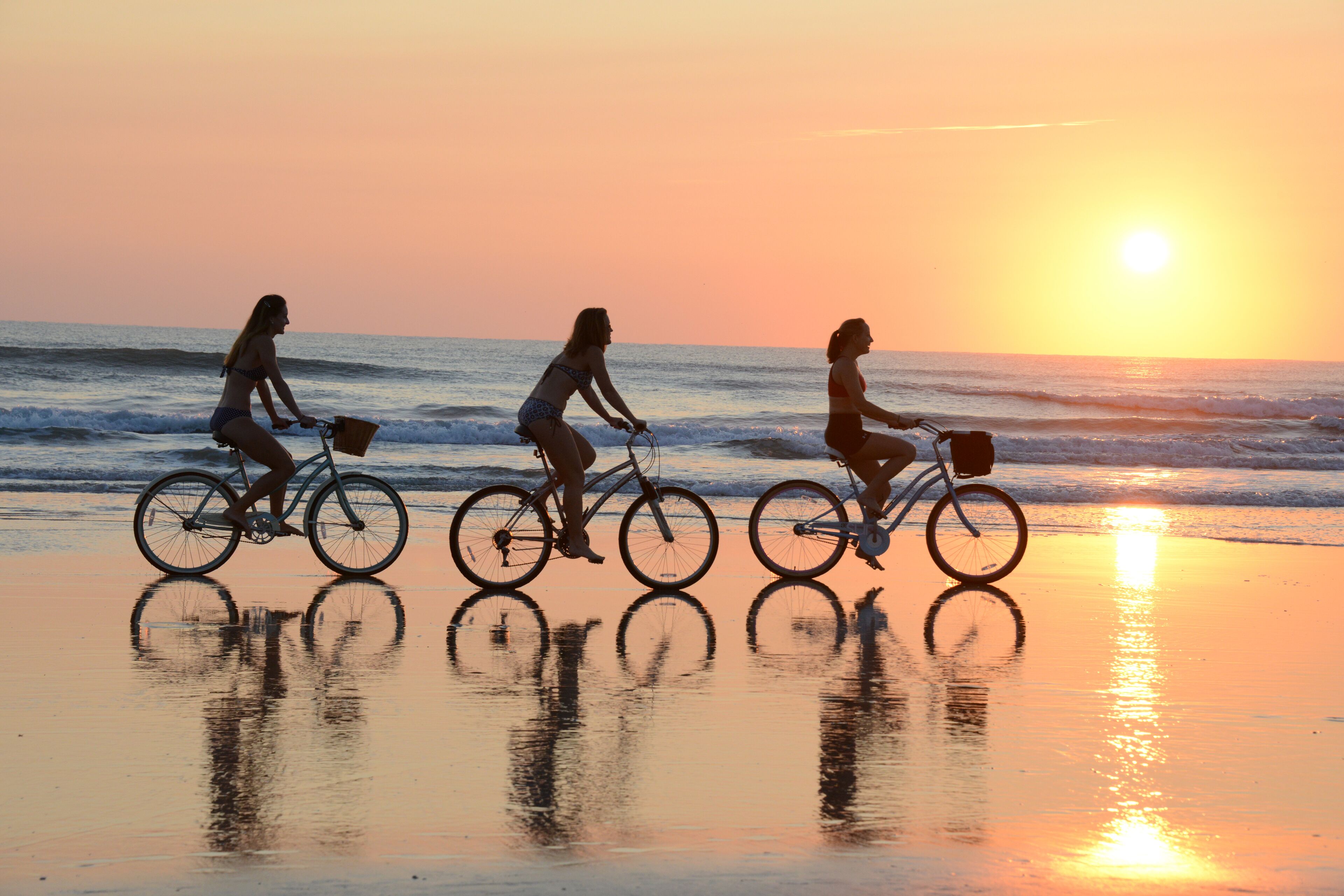 Three people riding bicycles on a beach at sunset.