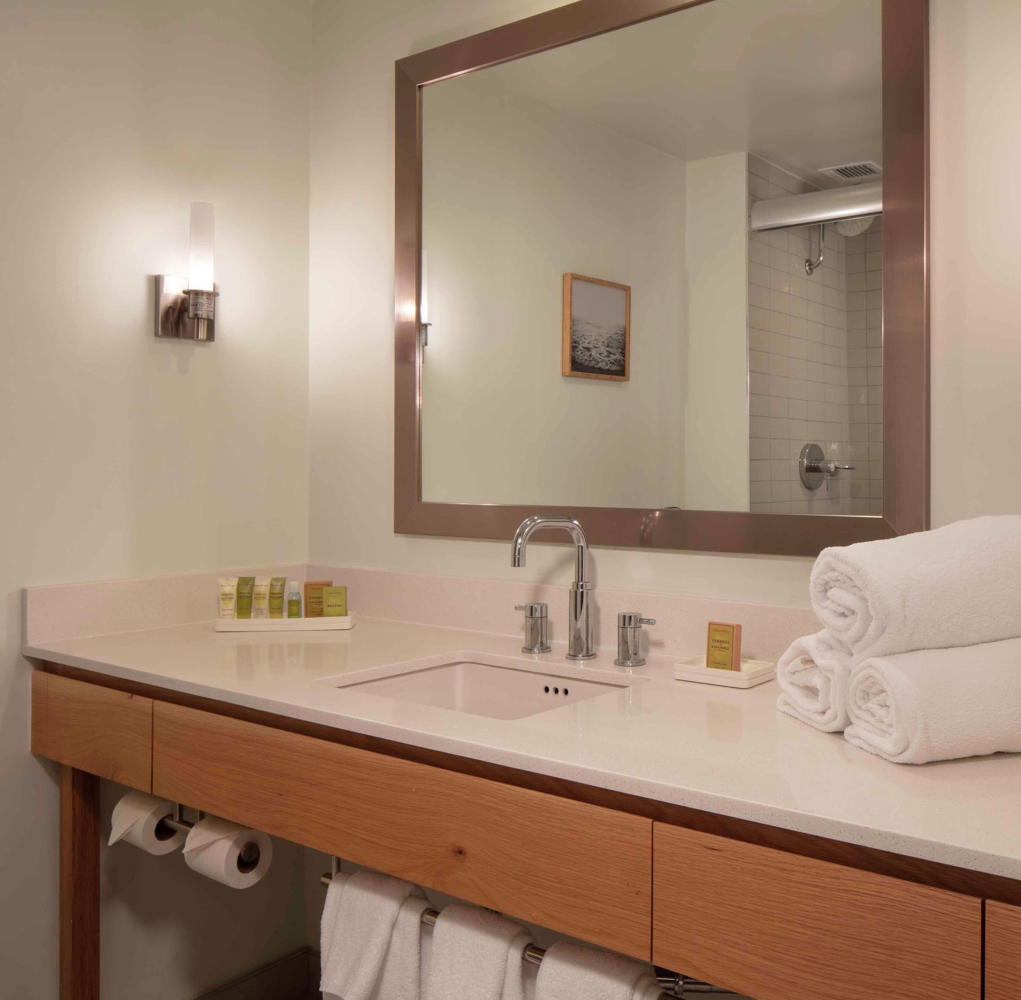 A hotel bathroom with a large mirror, two sinks, a towel rack, and a toilet paper dispenser.