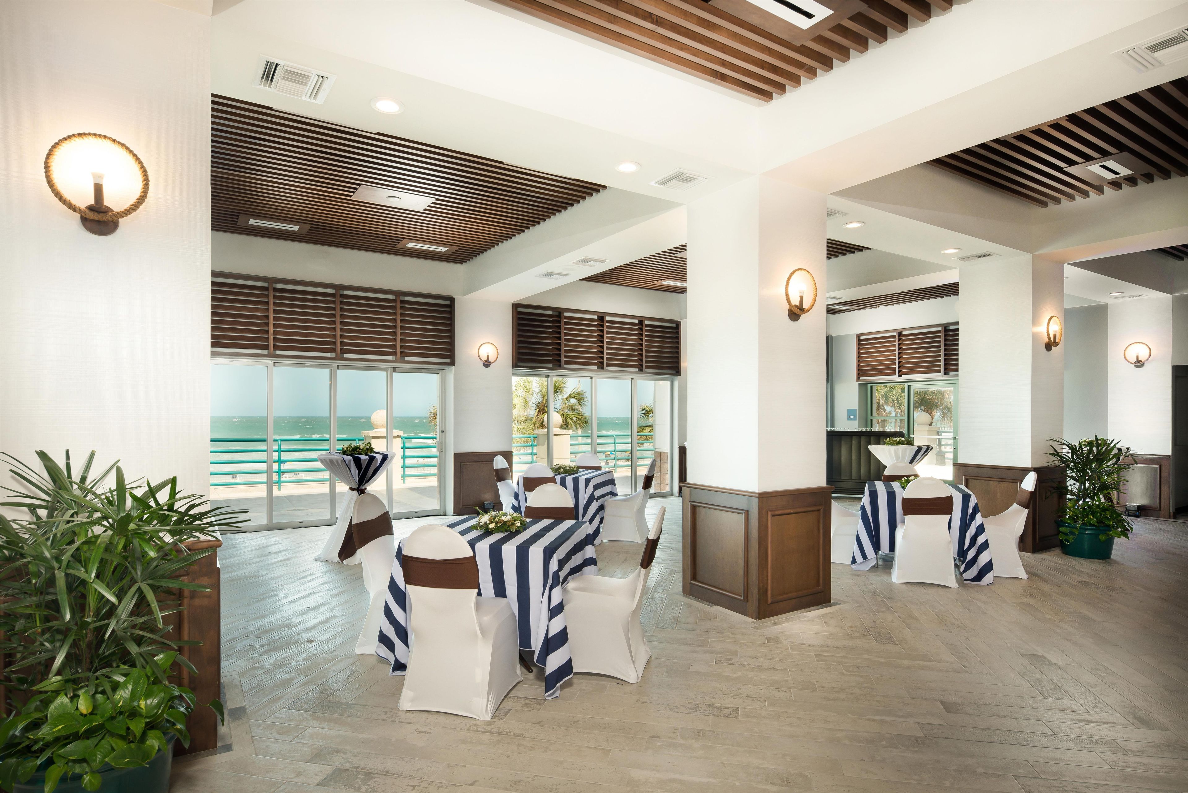 A spacious indoor area with multiple tables and chairs, large windows with ocean views, and a modern ceiling design.