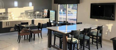 A modern dining area with a glass-top table, black chairs, and a kitchen area with a refrigerator and coffee machine.