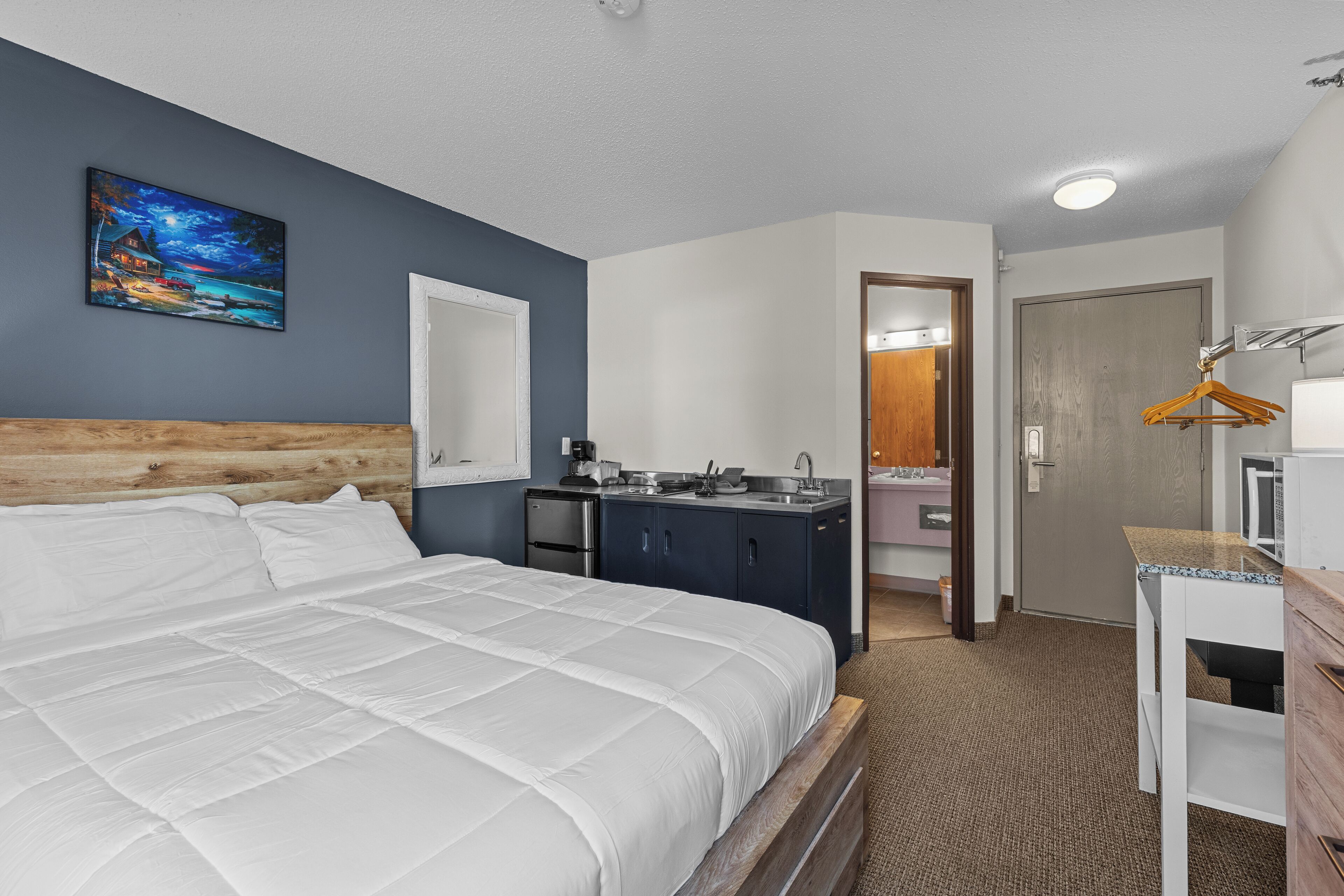 A hotel room with a large bed, a small kitchenette, a bathroom with a mirror, and a wooden headboard.