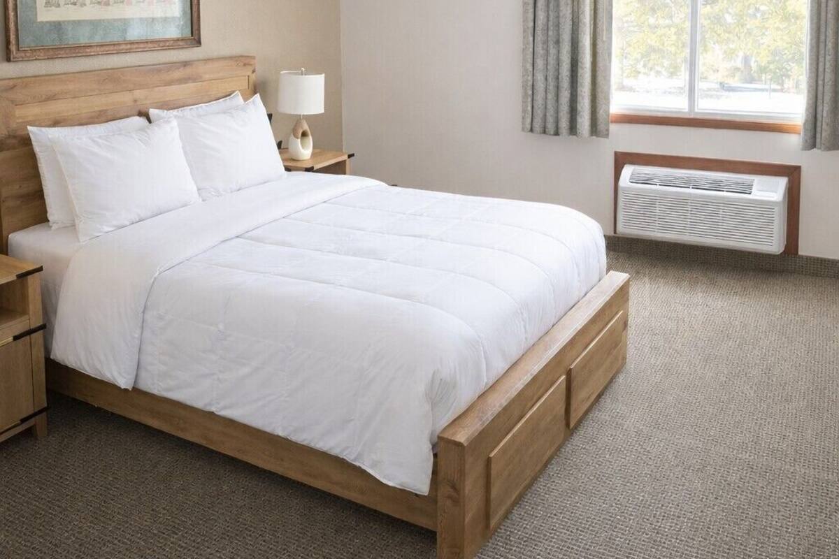 A neatly made bed with white linens, a wooden headboard, a bedside table with a lamp, a window with curtains, and a wall-mounted air conditioner.