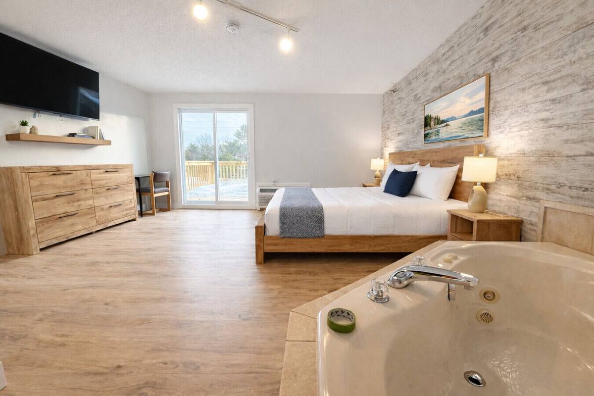 A spacious bedroom with a large bed, a wooden dresser, a TV, a bathtub, and a balcony with a view.