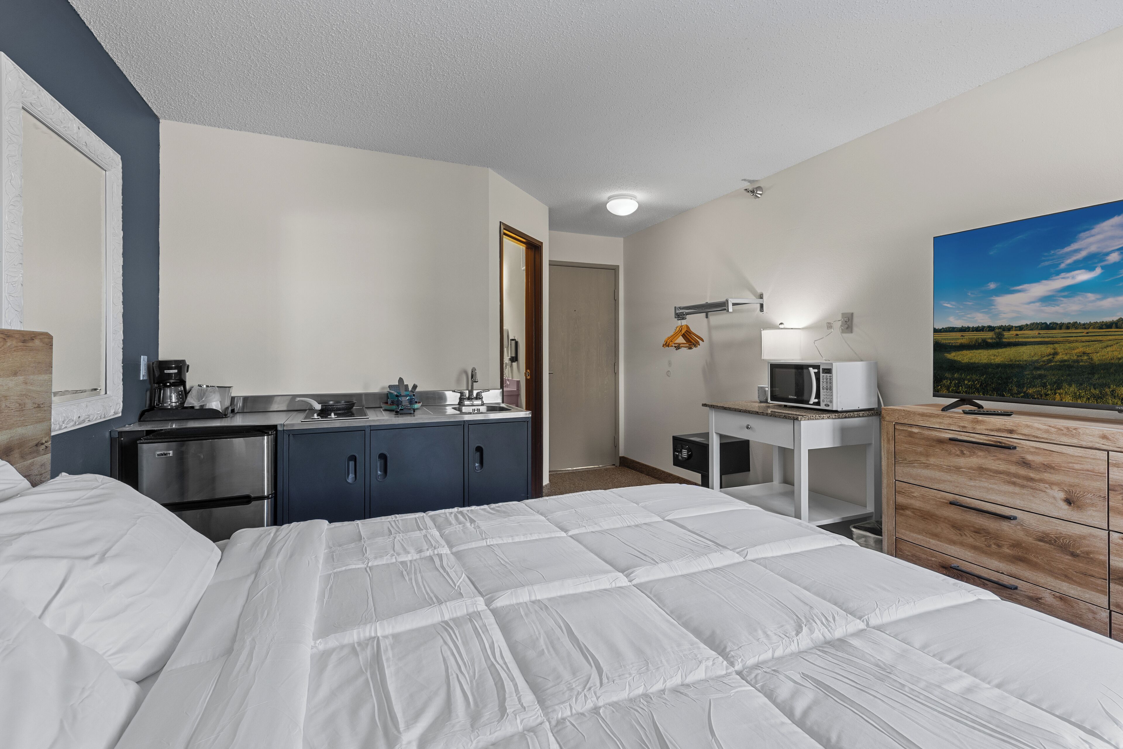 A hotel room with a bed, a kitchenette, a desk, and a painting on the wall.