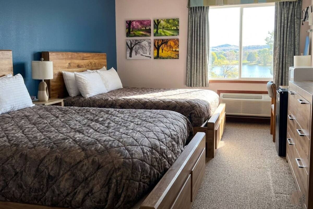 A hotel room with two beds, a wooden headboard, a nightstand with a lamp, a dresser, and a window with a view of a lake.