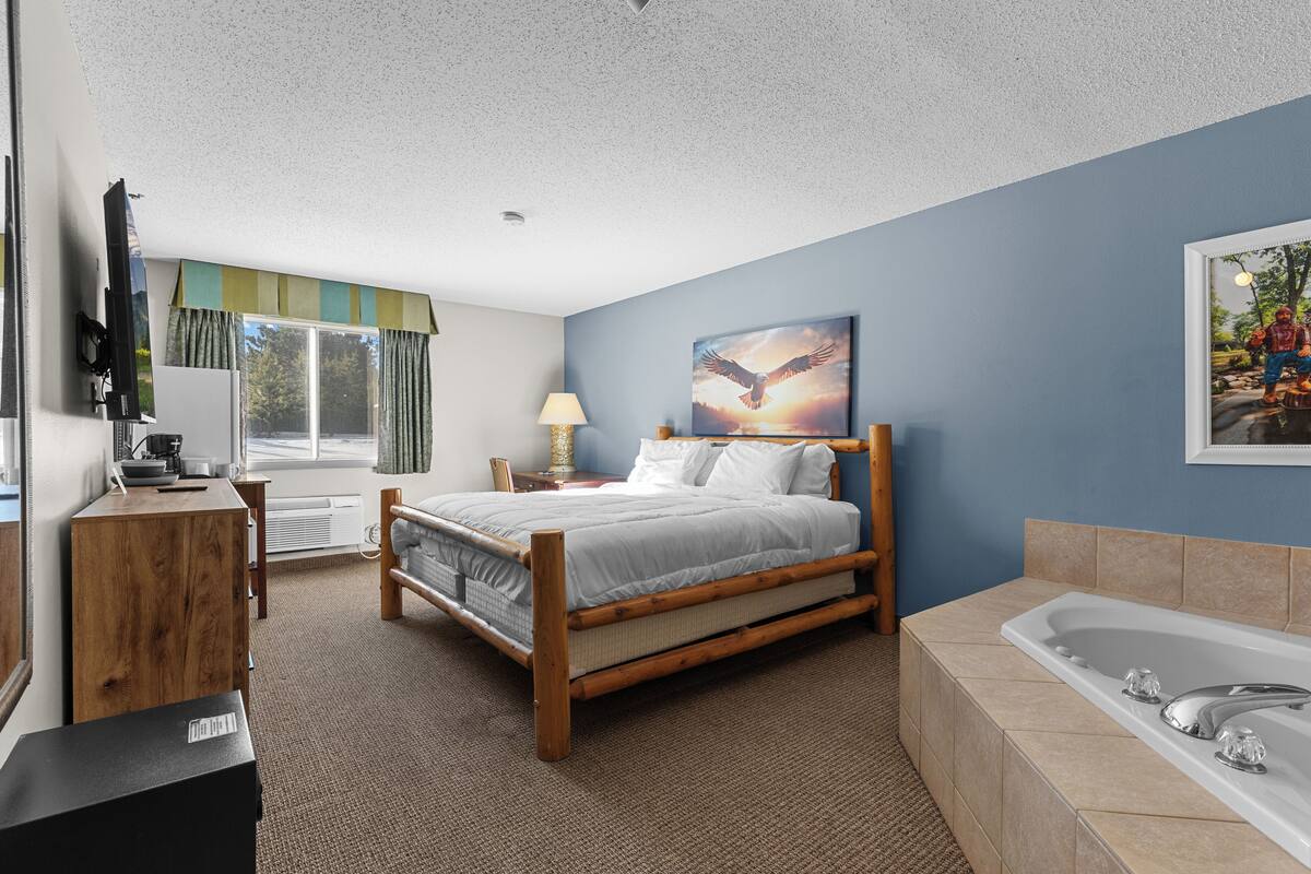 A bedroom with a bed, a wooden dresser, a desk, a lamp, a painting, and a bathtub.