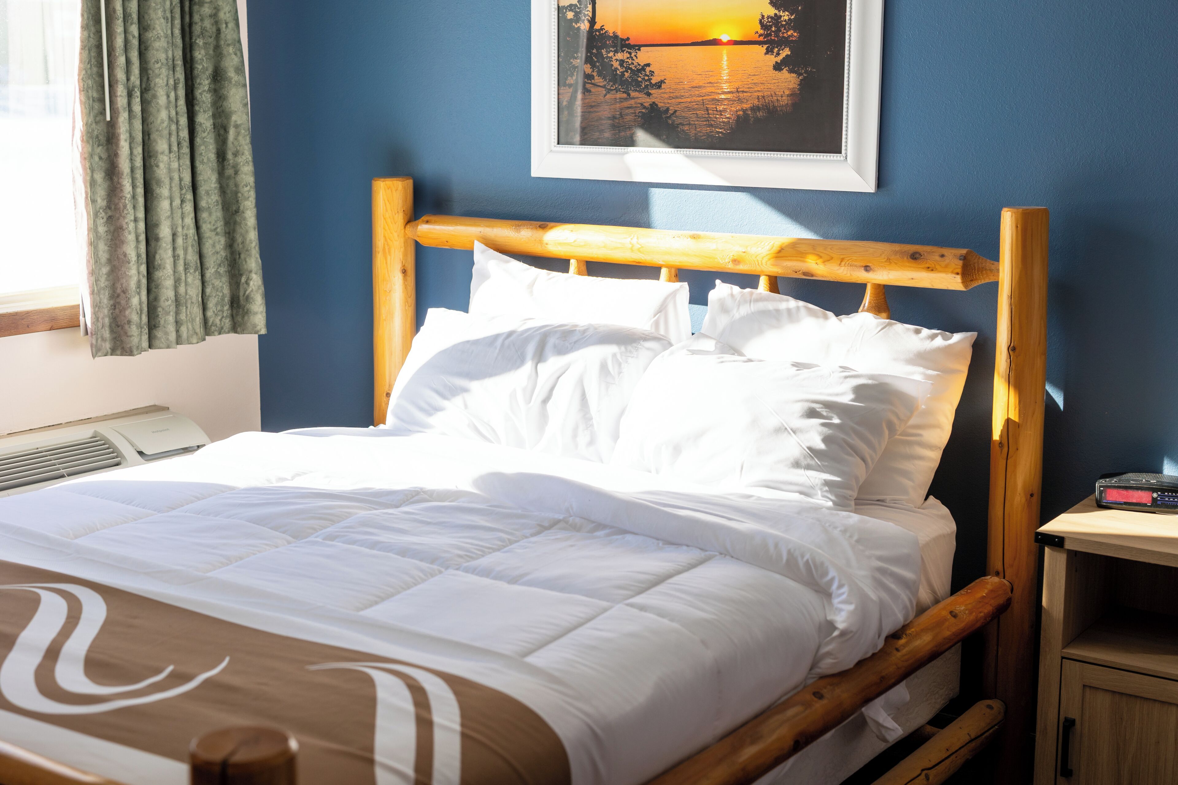 A neatly made bed with white linens, a wooden headboard, a bedside table with a clock, and a framed picture of a sunset on the wall.