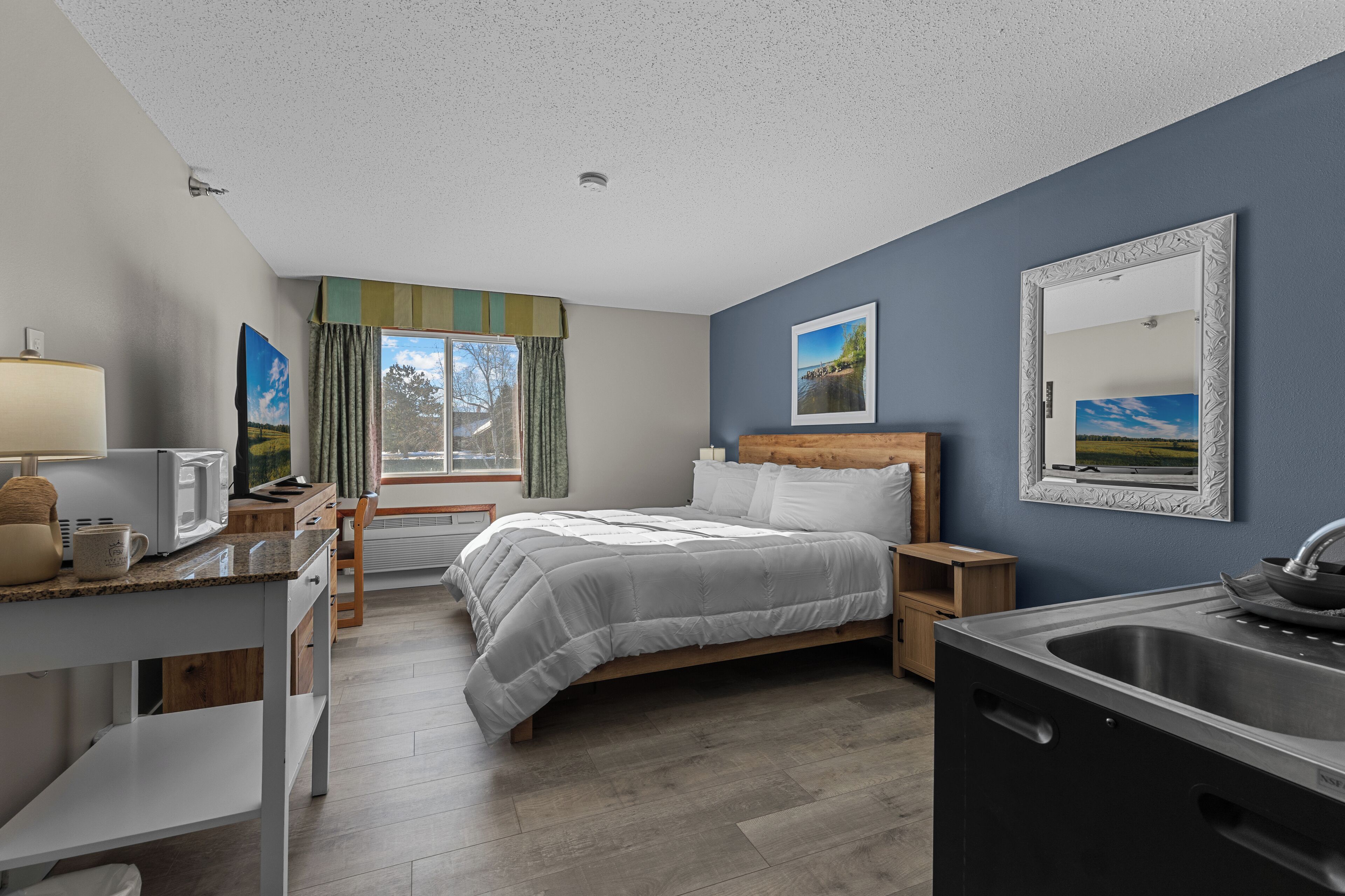 A hotel room with a bed, a desk with a lamp, a microwave, a sink, and a window with curtains.