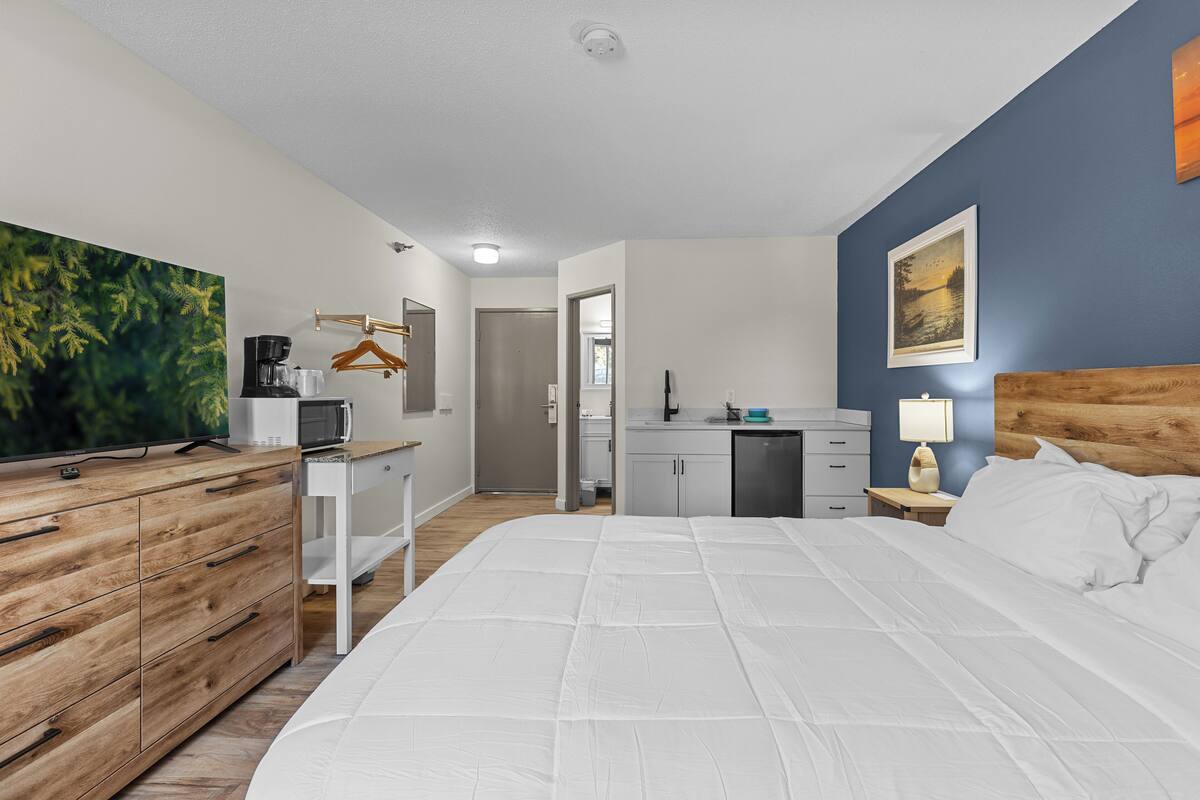 A hotel room with a bed, a desk, a dresser, a microwave, a coffee maker, a TV, and a painting on the wall.