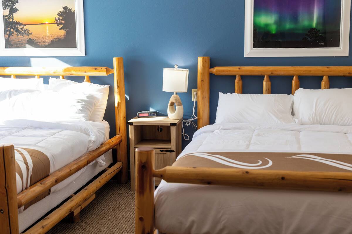 A hotel room with two wooden beds, a nightstand with a lamp, and framed pictures on the wall.