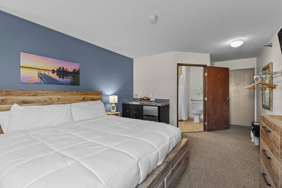 A hotel room with a bed, a painting of a sunset, a wooden headboard, a desk with a lamp, a bathroom with a toilet, and a wooden dresser.