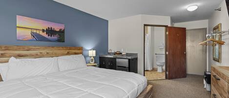 Habitación de hotel con cama, un cuadro de un atardecer, cabecero de madera, escritorio con lámpara, baño con inodoro y cómoda de madera.