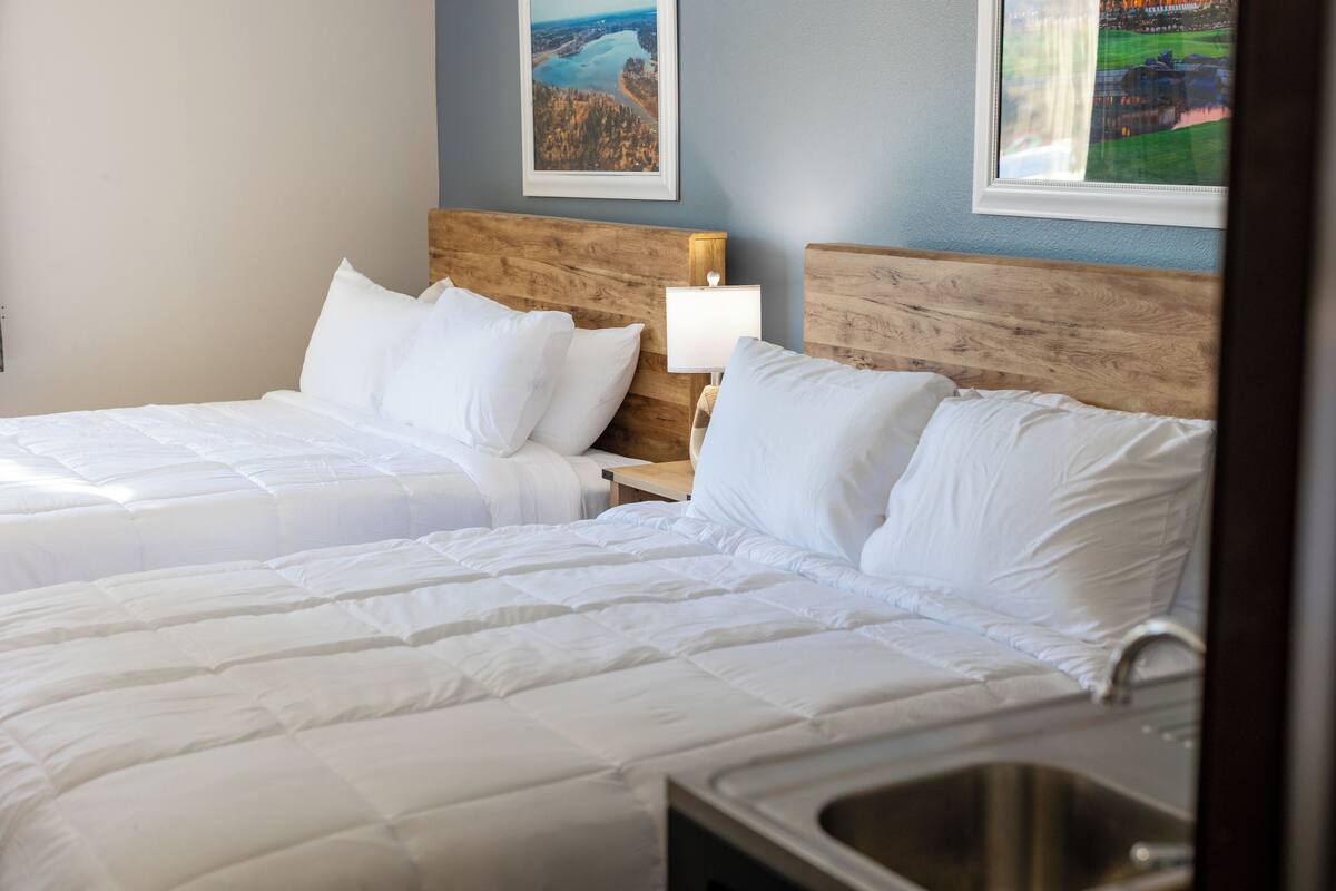 A hotel room with two beds, a wooden headboard, a nightstand with a lamp, and a window with a view of a landscape.