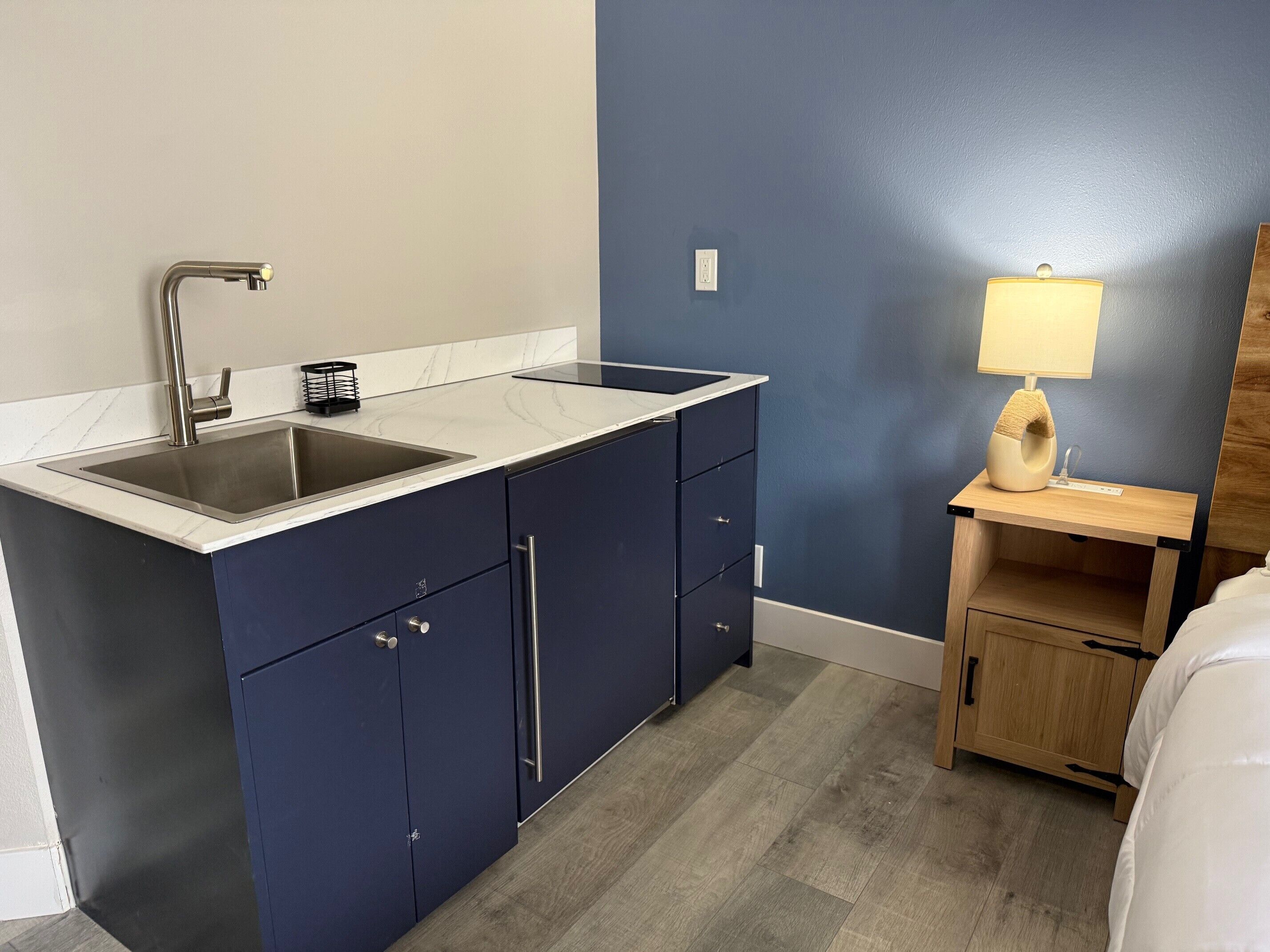 A compact kitchenette with a sink, cabinets, and a small bedside table with a lamp.