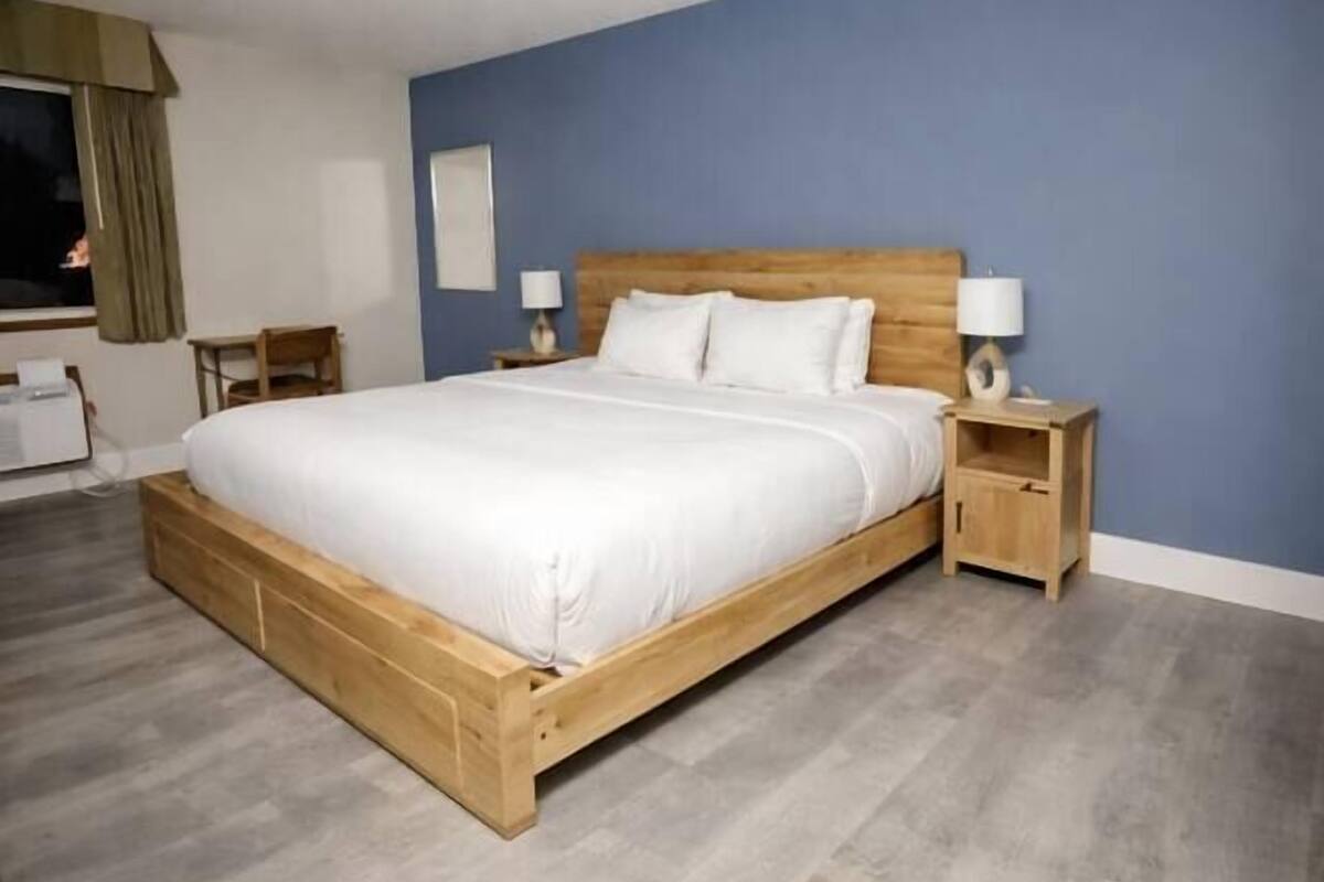 A hotel room with a wooden bed, two bedside tables with lamps, a window with curtains, and a desk with a chair.