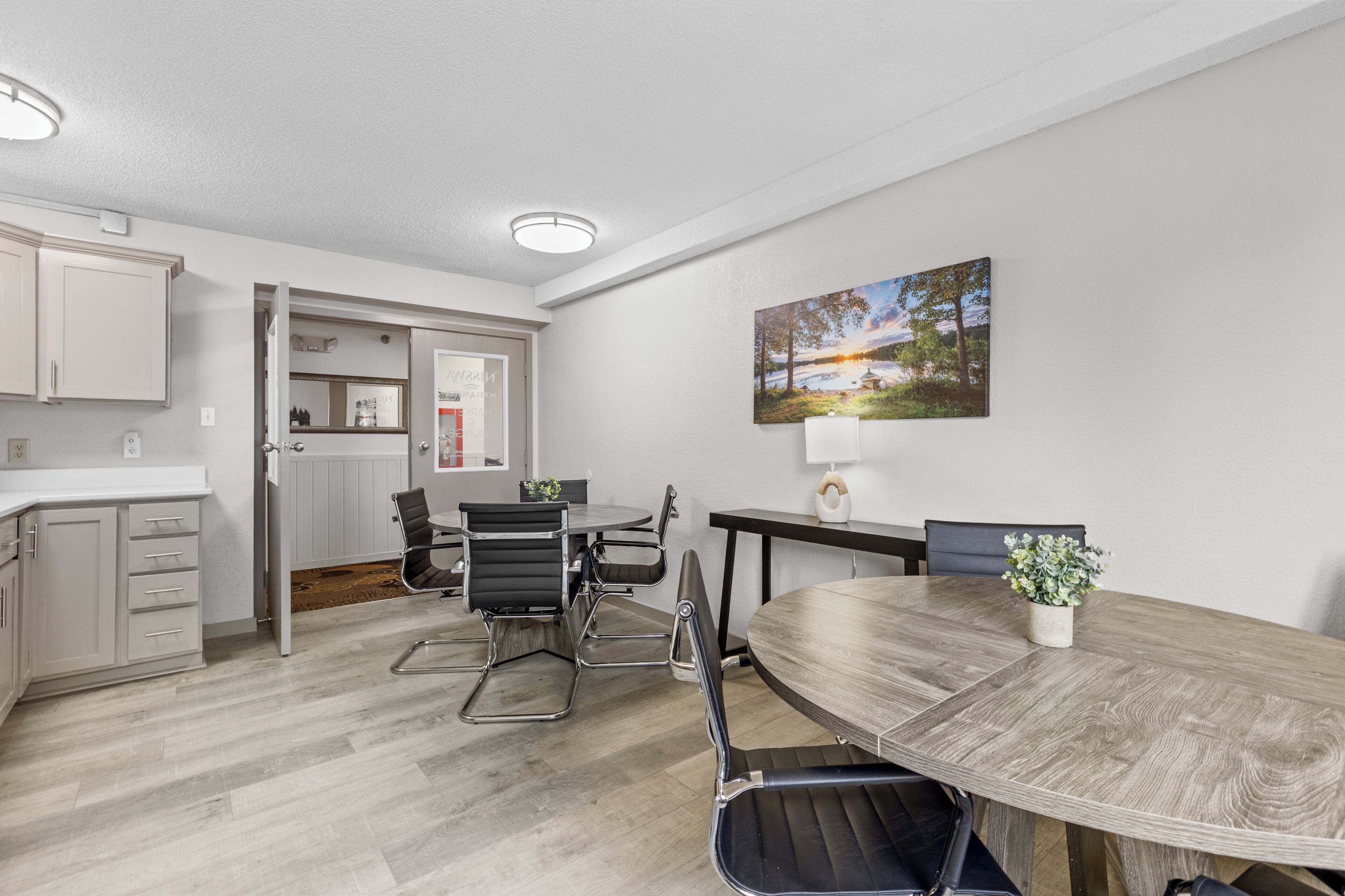 A modern kitchen with white cabinets, a dining area with a wooden table and black chairs, and a painting on the wall.