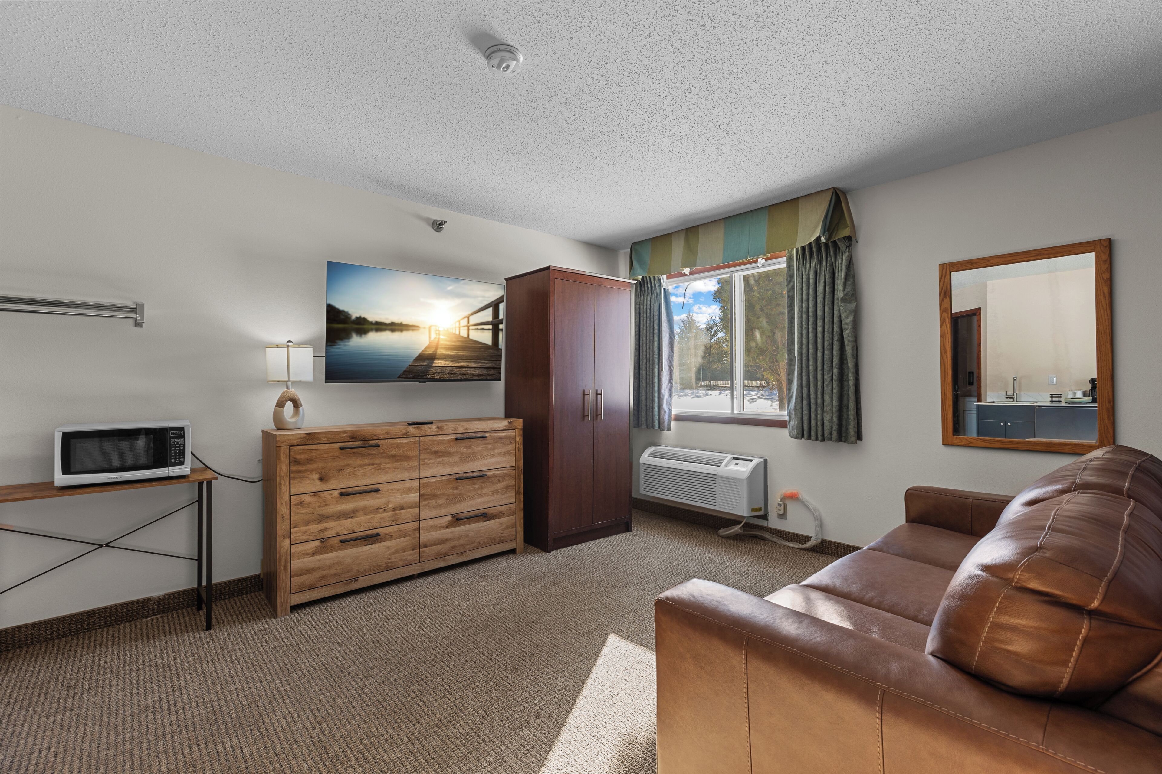 A room with a brown leather sofa, wooden cabinet, microwave, and a painting of a sunset over a bridge.