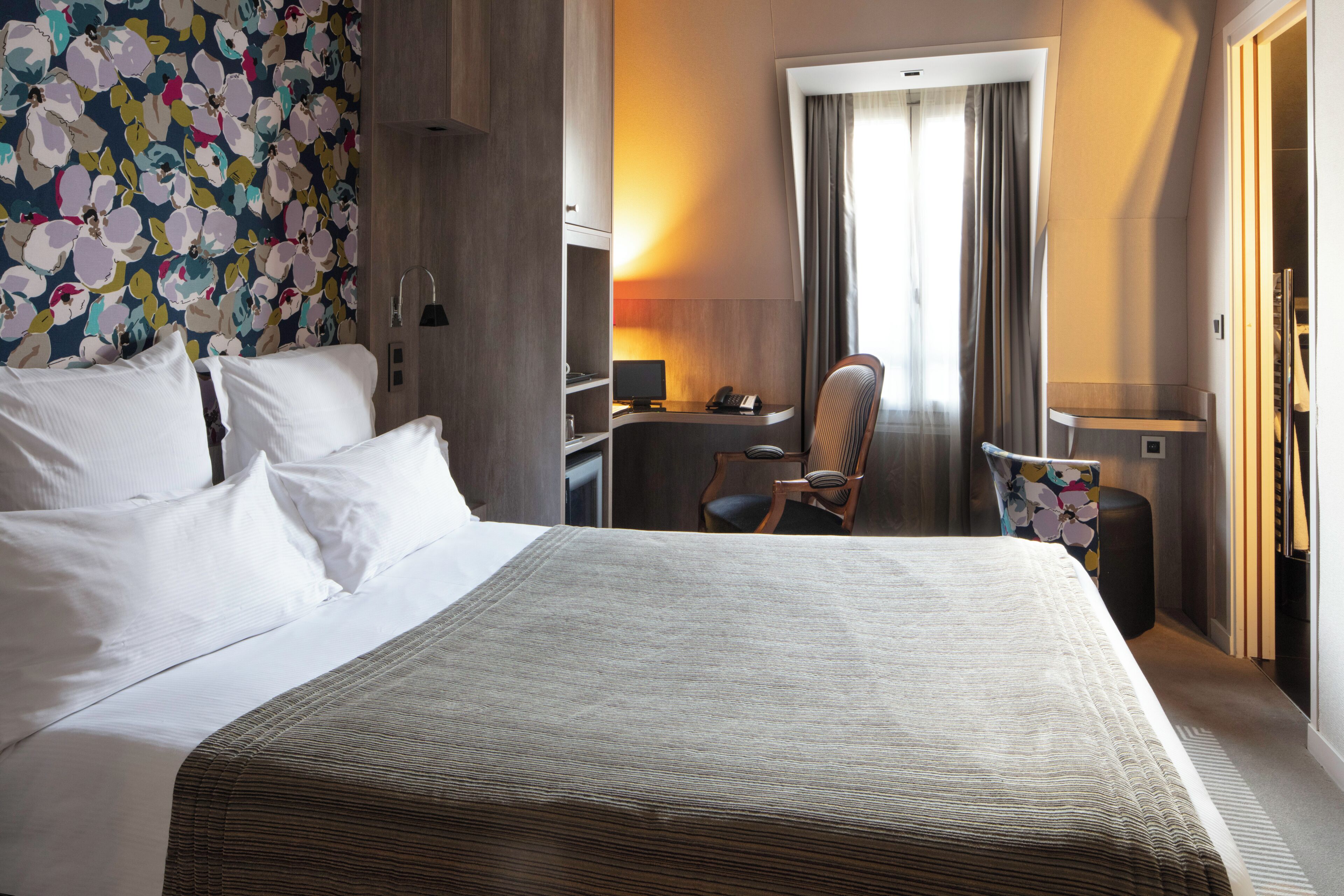 A hotel room with a bed, desk, chair, and a large window with curtains.