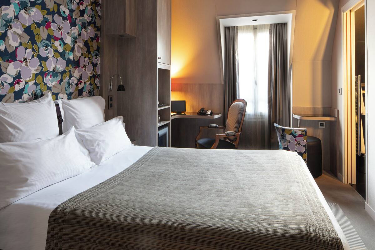 A hotel room with a bed, desk, chair, and a large window with curtains.