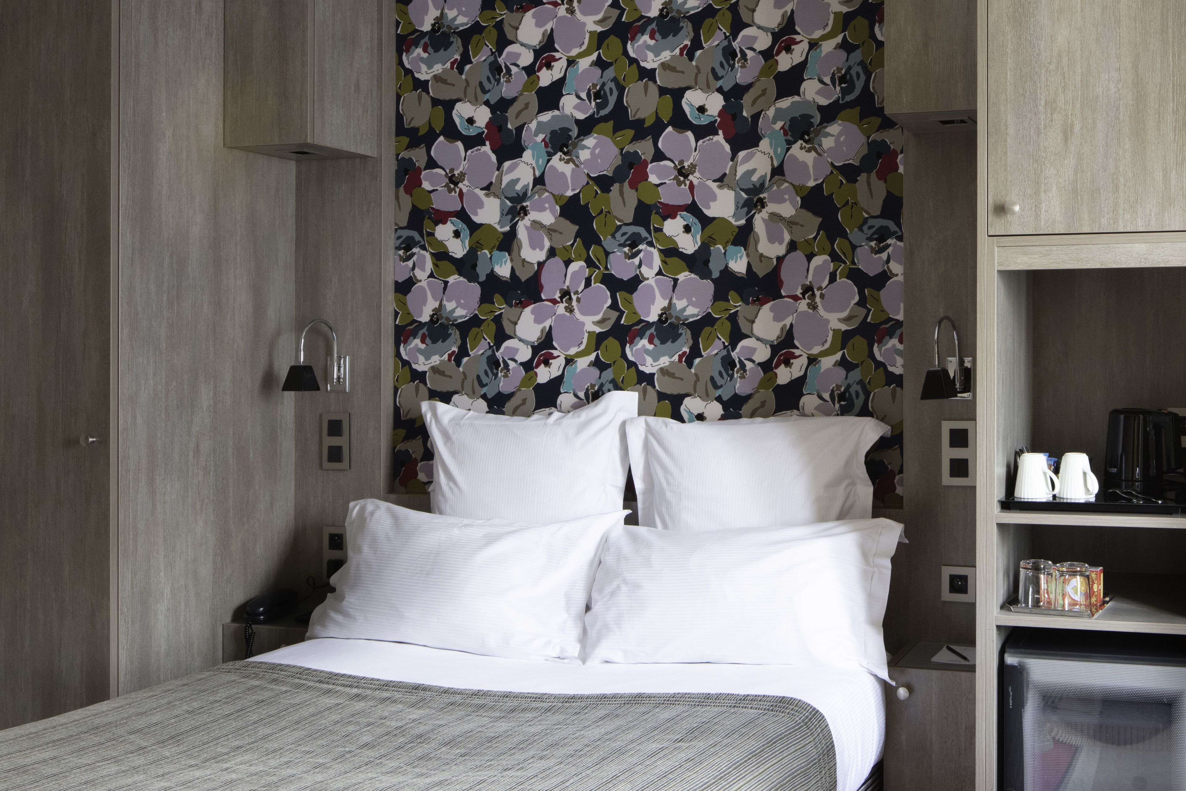 A hotel room with a bed, white pillows, a floral-patterned wall, a desk with a phone, and a shelf with cups and a kettle.