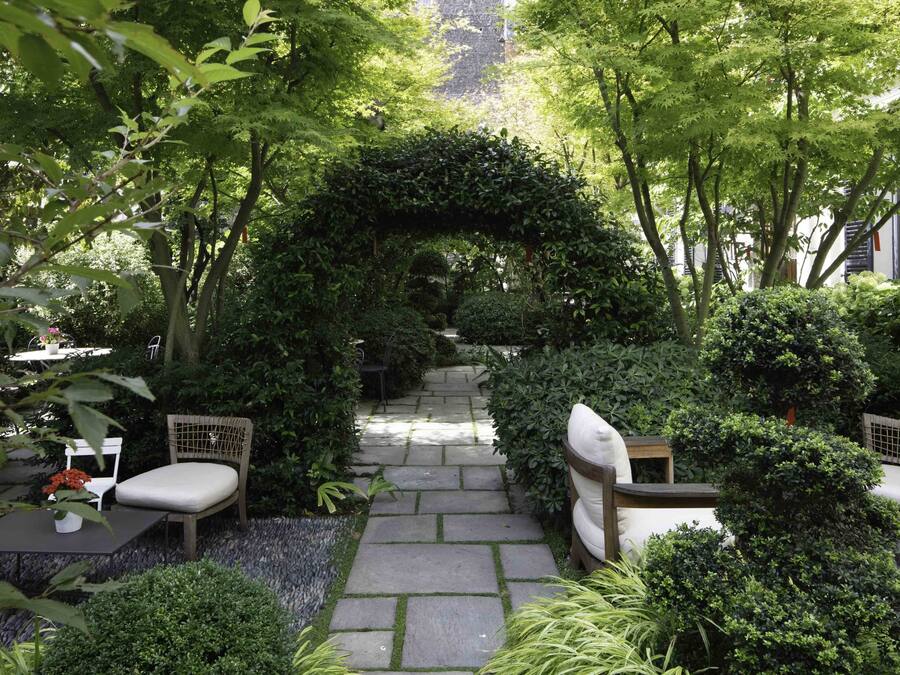 A garden pathway with a stone archway, surrounded by lush greenery and seating areas.