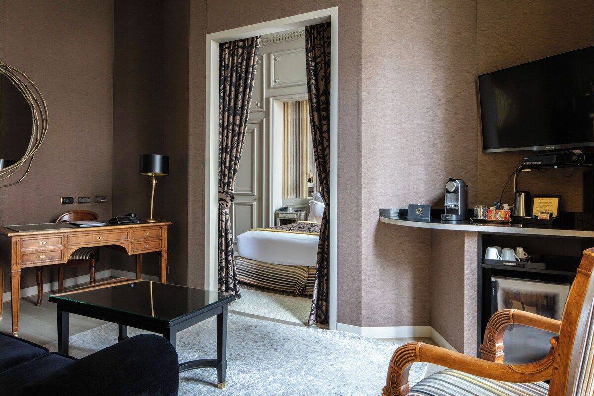 A hotel room with a desk, a chair, a bed, a television, and a coffee machine.