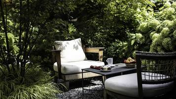 Un espace extérieur aménagé avec des sièges, comprenant un fauteuil confortable et une table, le tout entouré d’une végétation luxuriante et bordé d’un chemin en pierre.