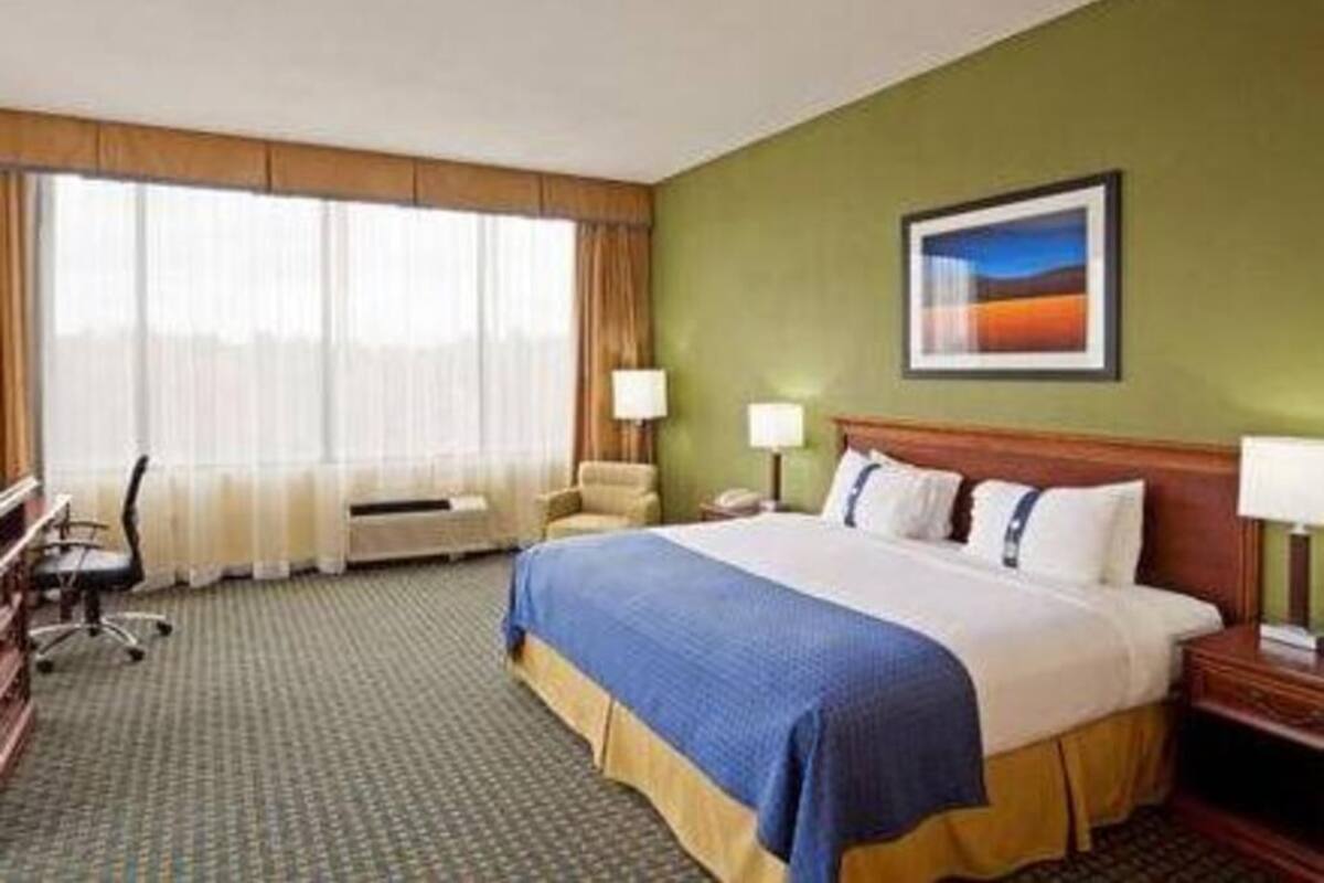 Habitación de hotel con cama, escritorio, silla, ventana con cortinas y un cuadro en la pared.