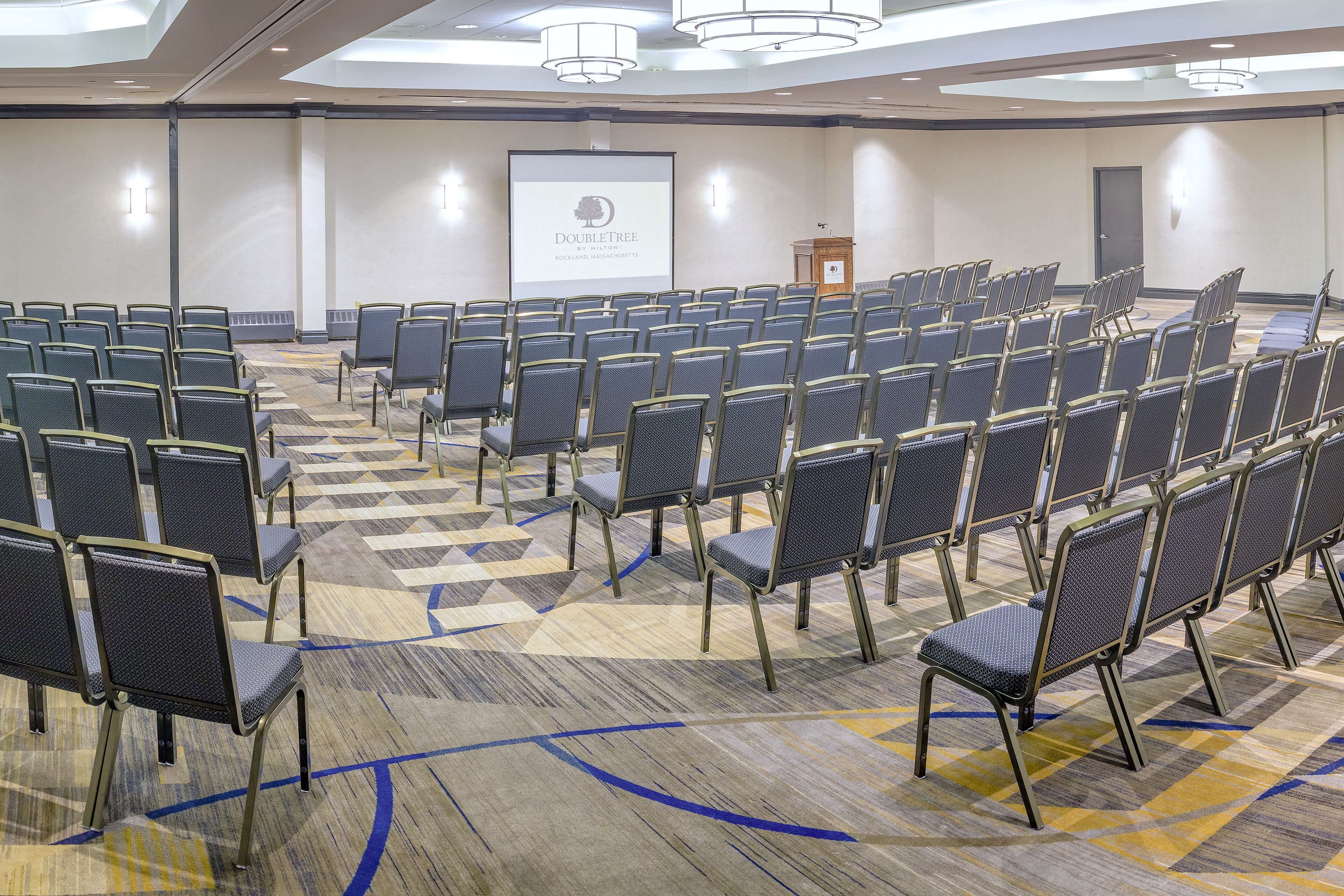 Een congreszaal met rijen stoelen, een spreekgestoelte en een presentatiescherm.