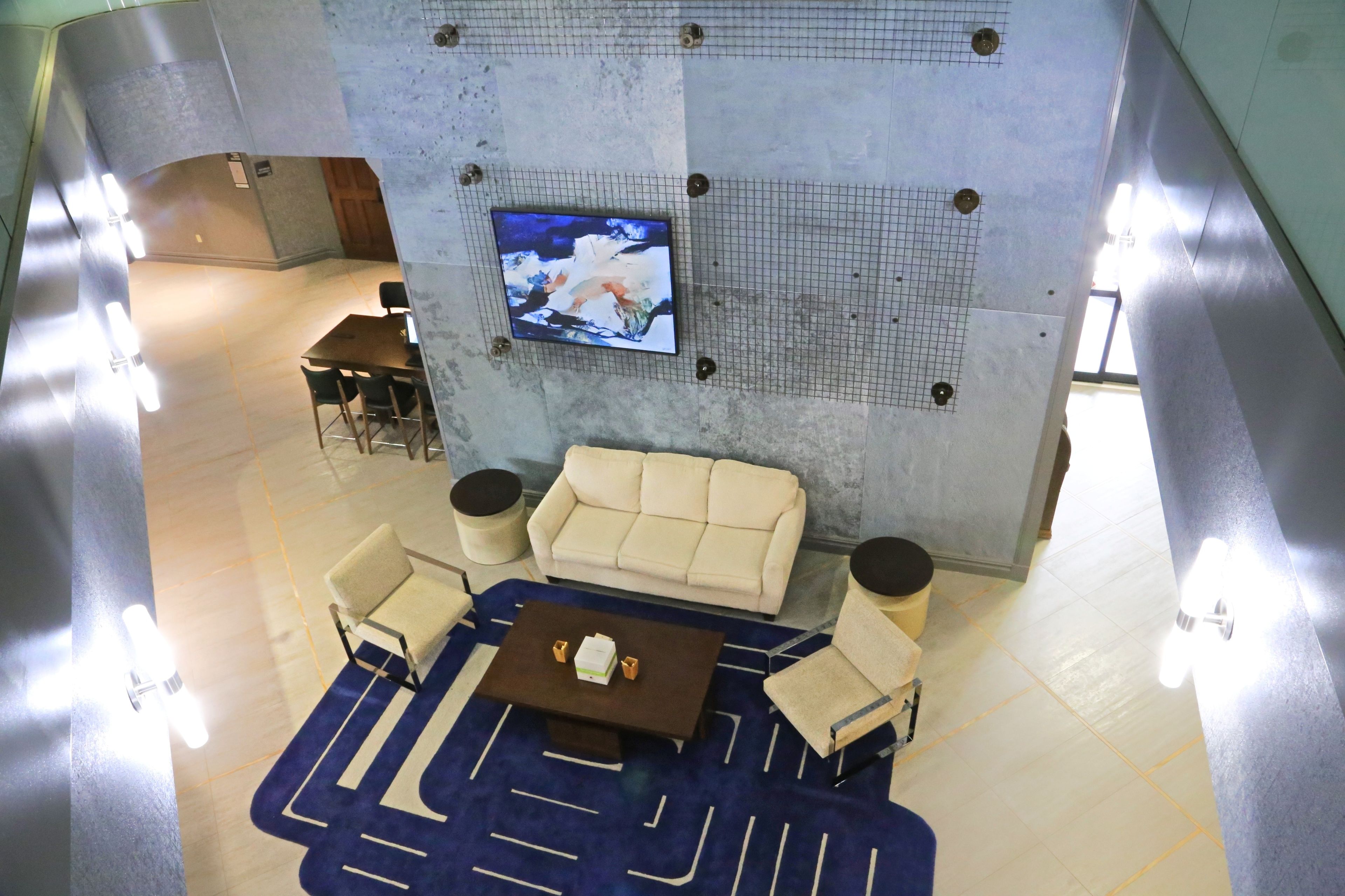 A hotel lobby with a seating area featuring a sofa, armchairs, and a coffee table. A television is mounted on the wall, and the floor is tiled.