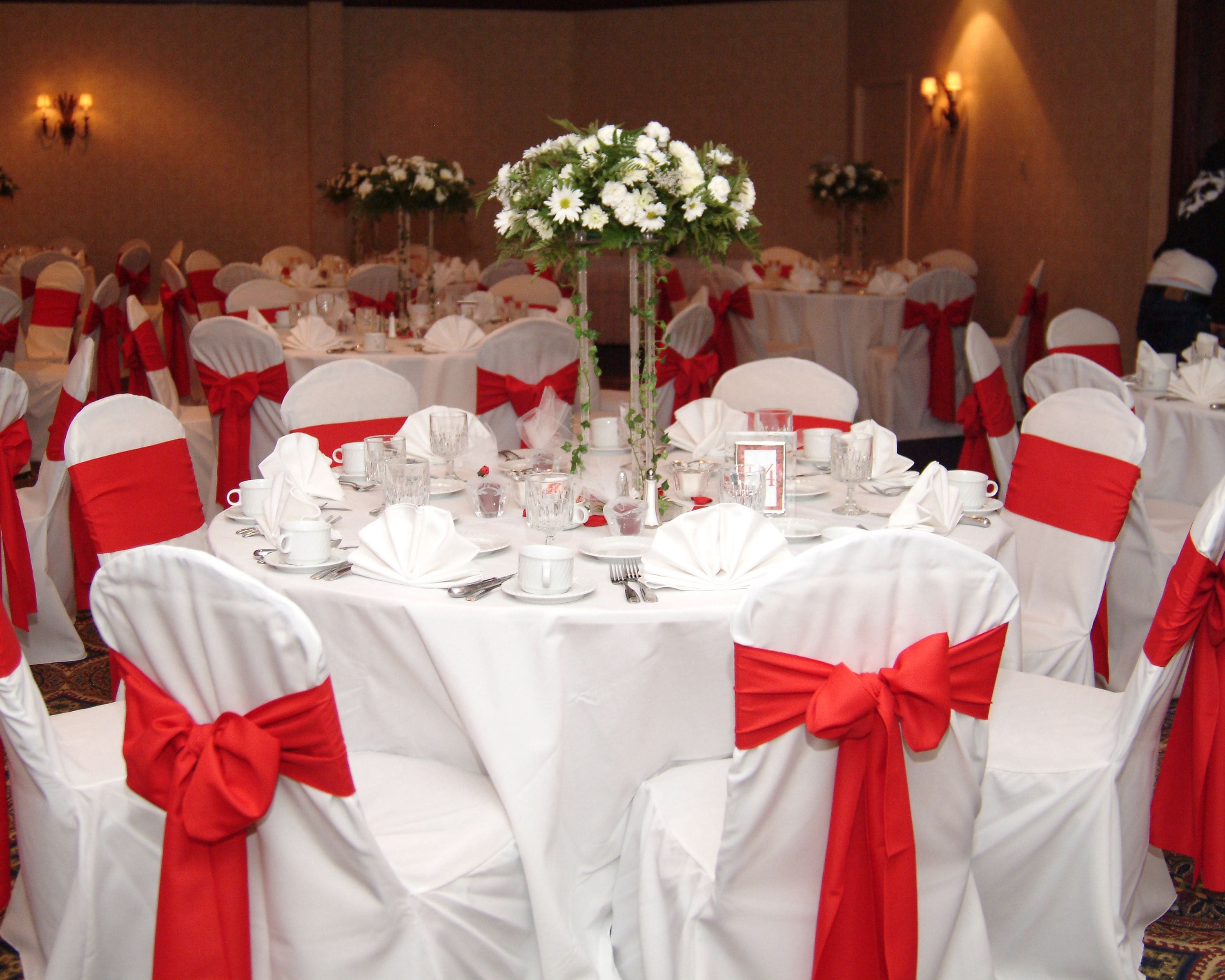 A banquet hall with round tables set for a formal event, each with white tablecloths and red sashes on the chairs.