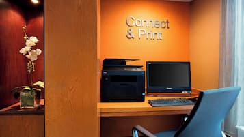 A hotel room with a desk featuring a Dell printer and an Apple monitor, a blue chair, and a sign that reads "Connect & Print".