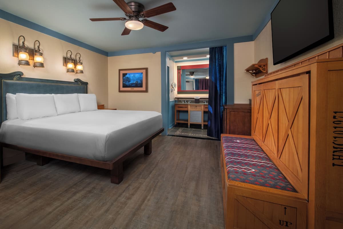 Habitación de hotel con cama, mesitas de noche de madera, ventilador de techo, luces fijadas a la pared, un cuadro enmarcado y un televisor.