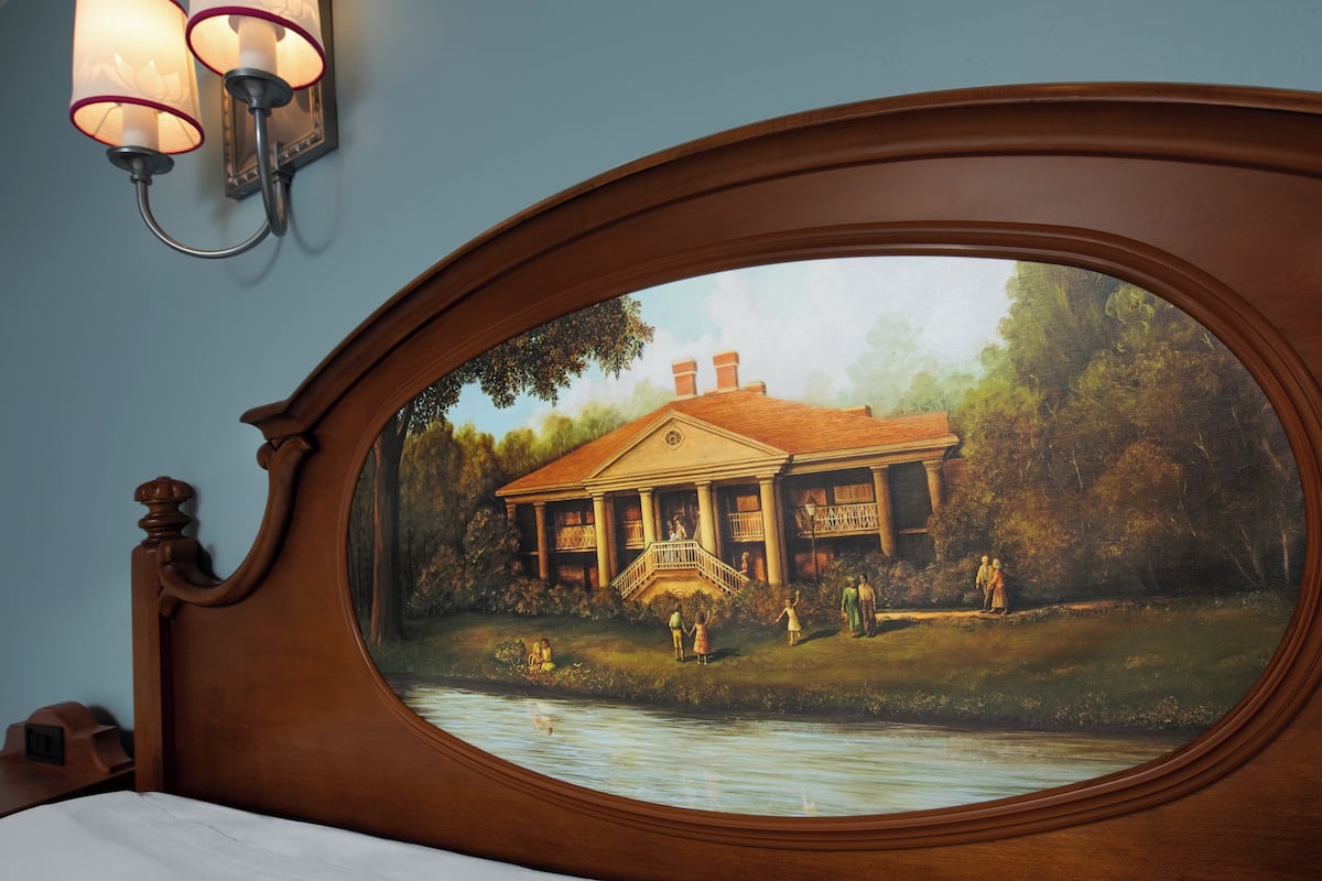 Una habitación con un espejo ovalado grande que refleja un cuadro de una casa junto a un río.