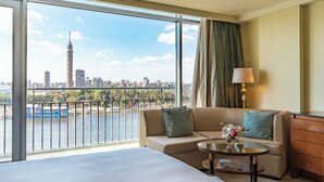 A hotel room with a large window offering a city view, a bed, a sofa, a small table with a lamp, and a dresser. - The Nile Ritz-Carlton, Cairo (Cairo)