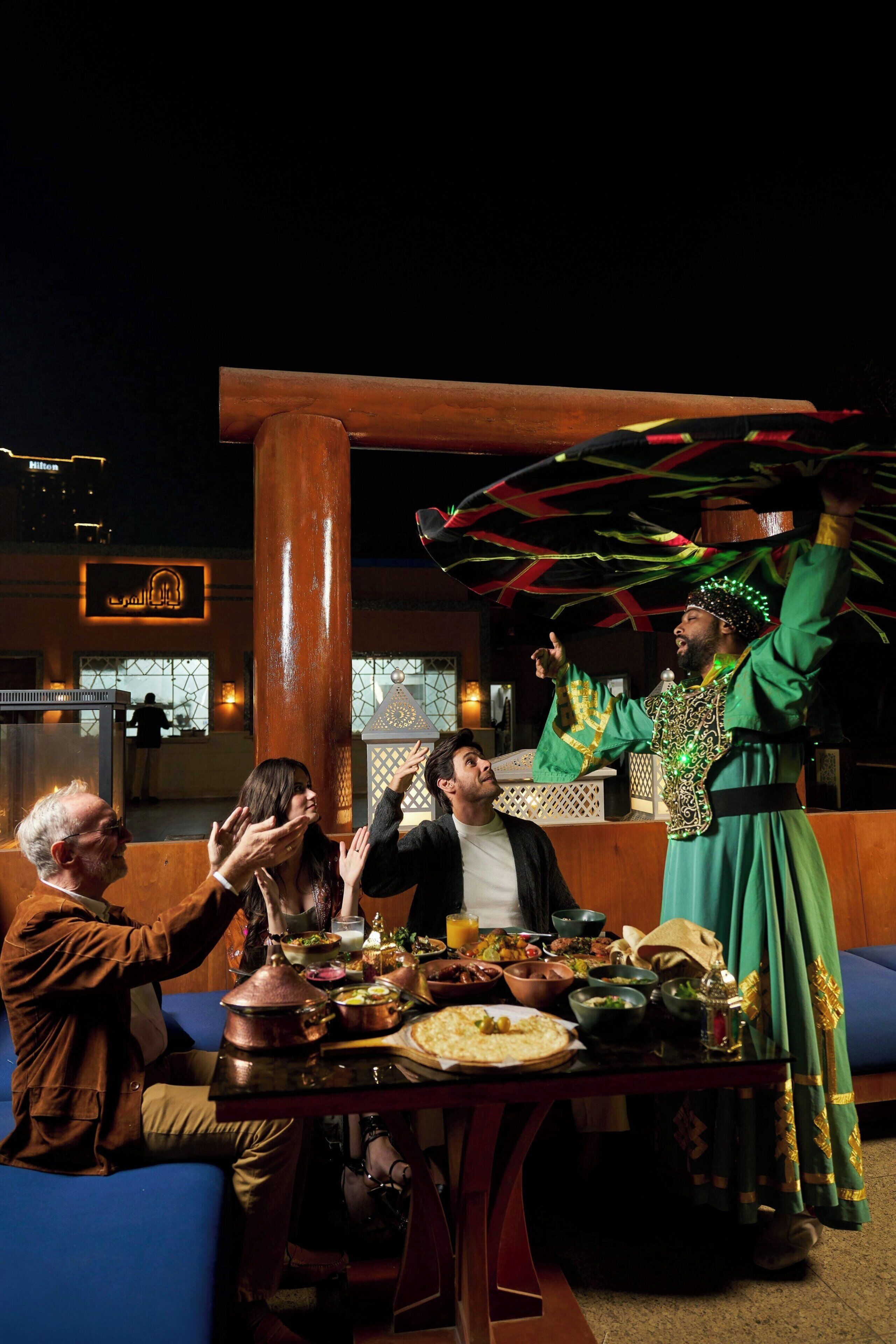 Um grupo de pessoas jantando ao ar livre à noite, com uma mesa posta para refeição, um pilar grande e um edifício com letreiros iluminados ao fundo.