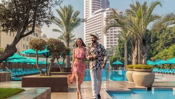 A couple walking by a pool with palm trees and a high-rise building in the background.