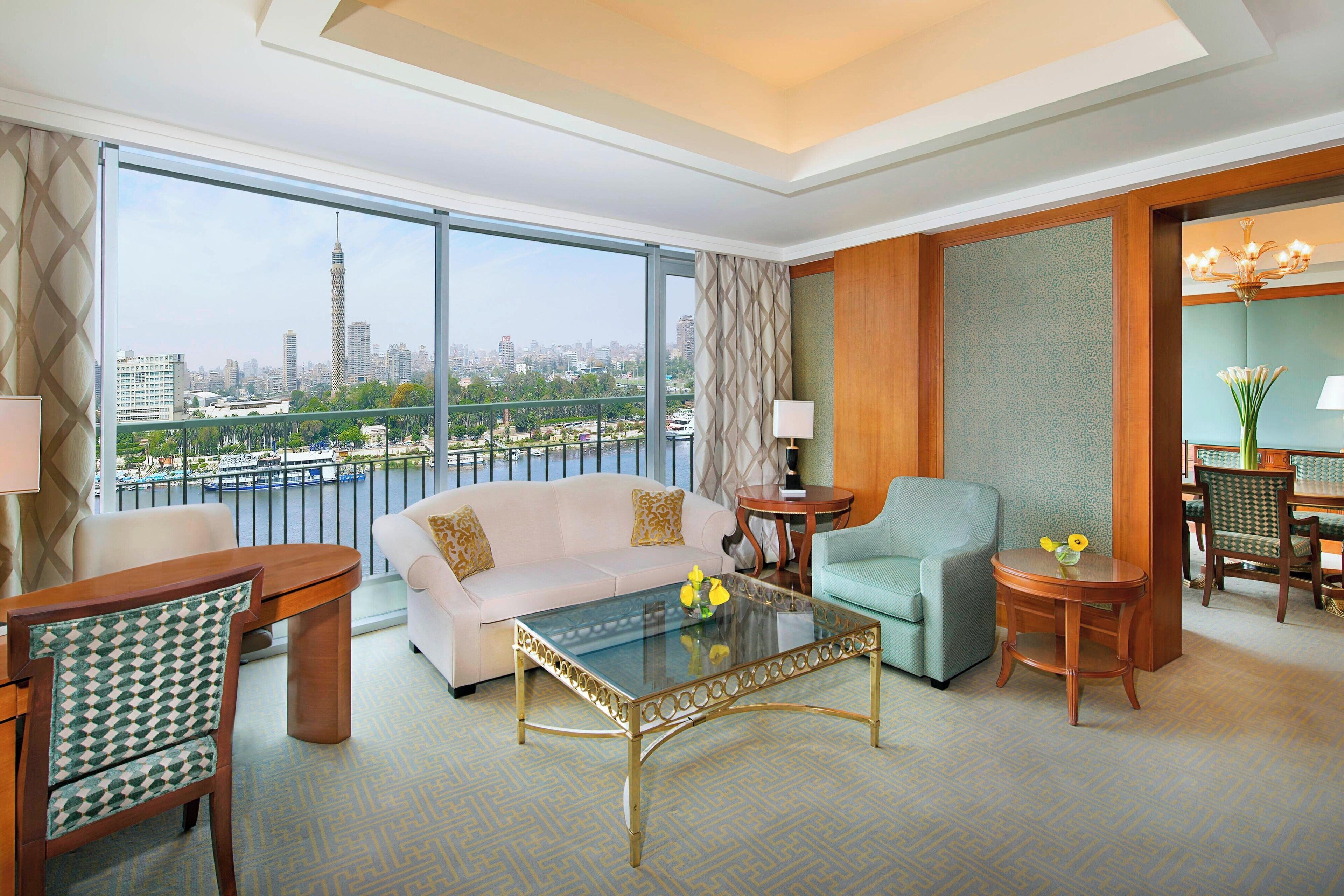 A modern hotel room with a large balcony, a glass coffee table, a sofa, a chair, and a dining area.