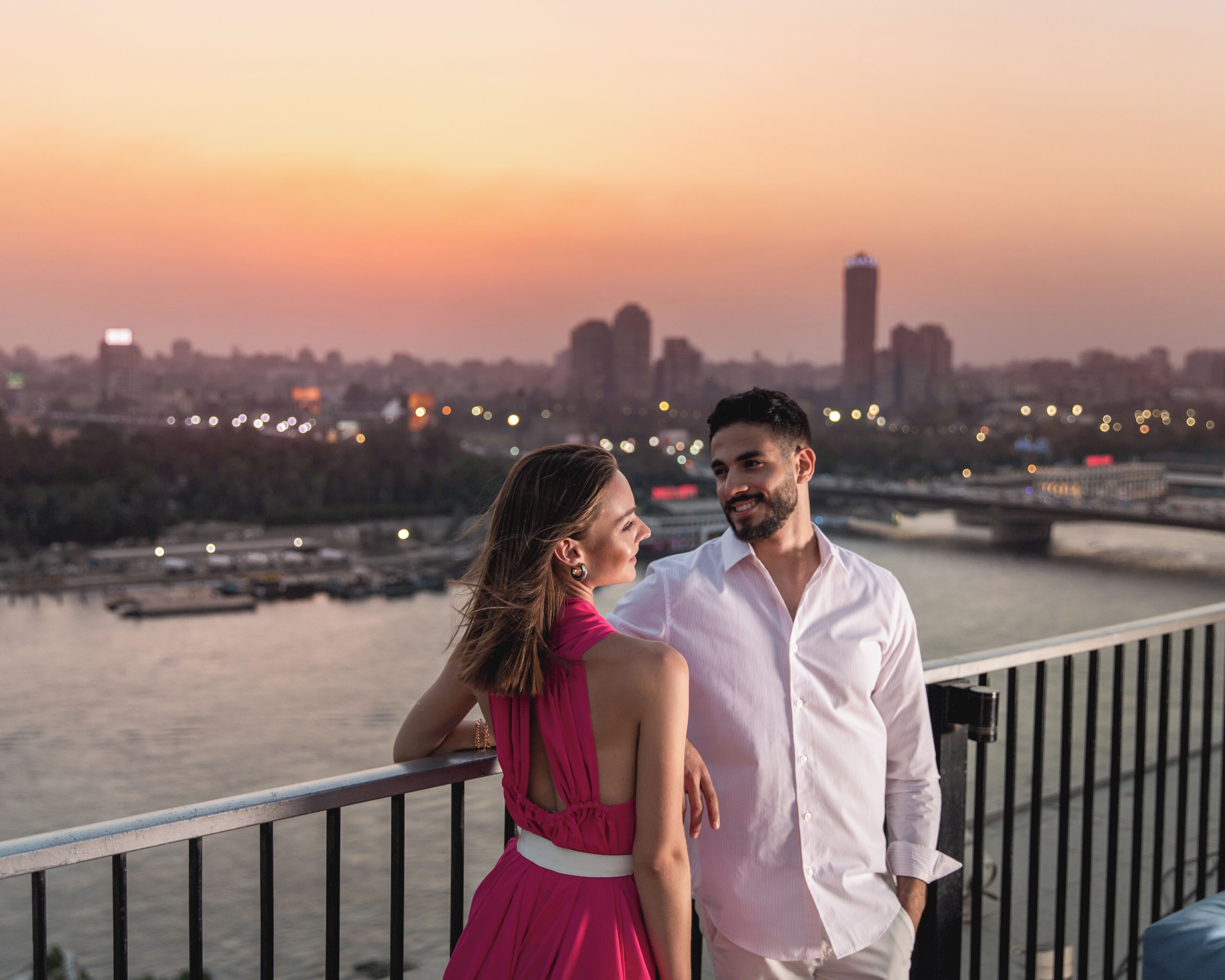 Una pareja parada en un balcón con vista a un río y el perfil urbano de una ciudad al fondo.