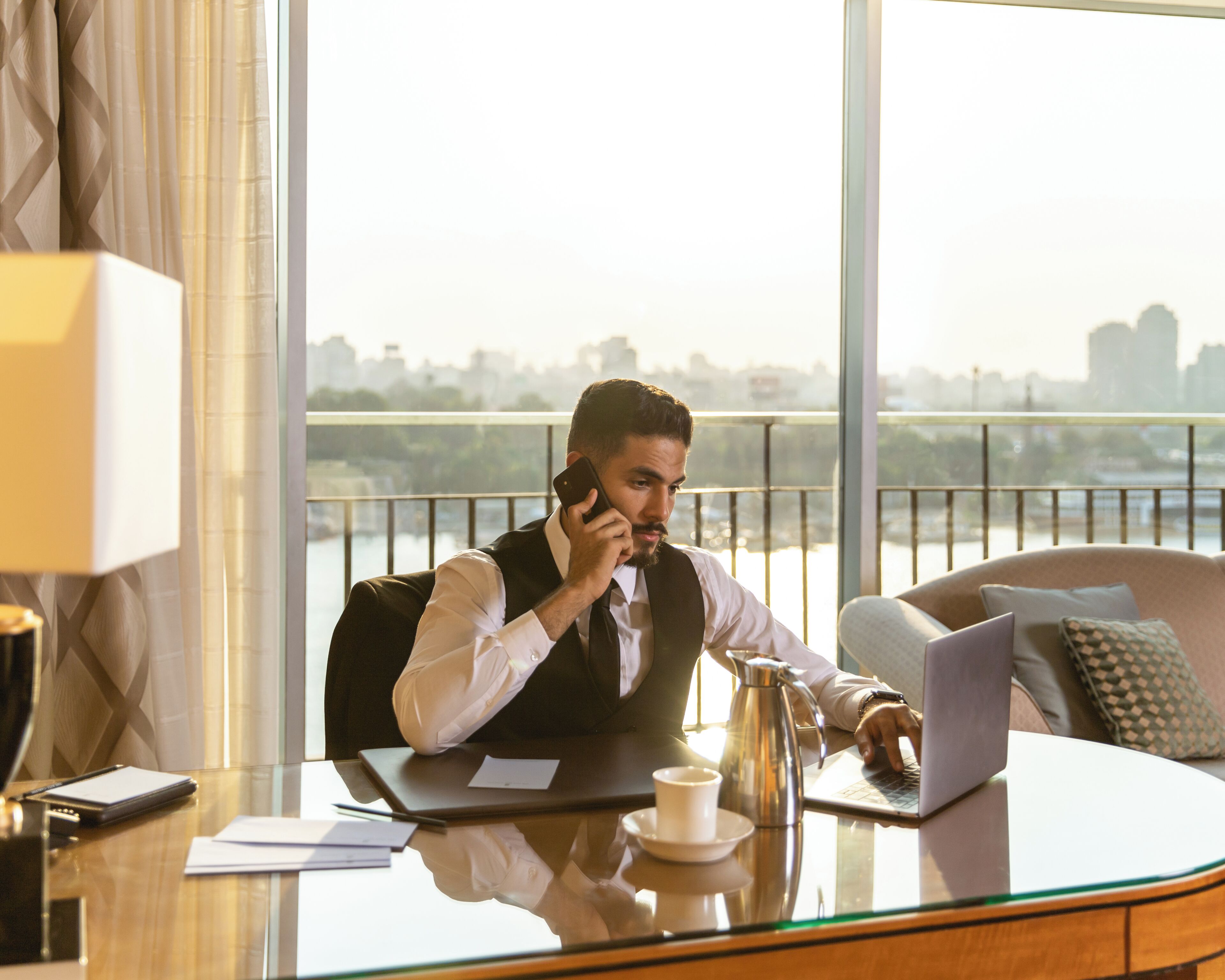 A man in a suit sitting at a desk with a laptop, talking on a phone, in a room with a city view.
