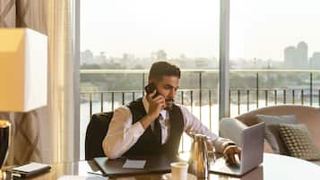 A man in a suit sitting at a desk with a laptop, talking on a phone, in a room with a city view.