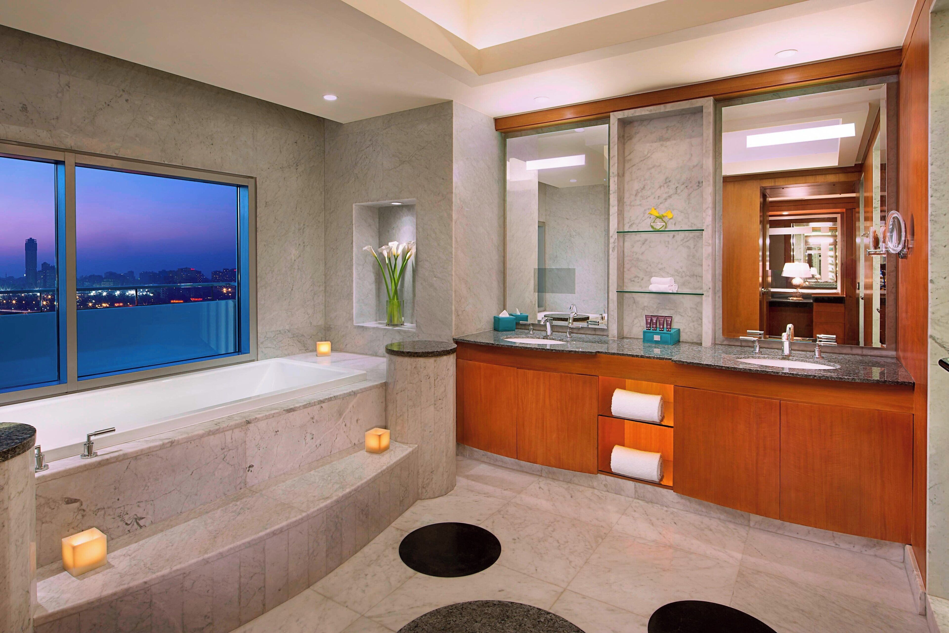 A modern bathroom with a large bathtub, a vanity with a mirror, and a view of the cityscape at dusk.